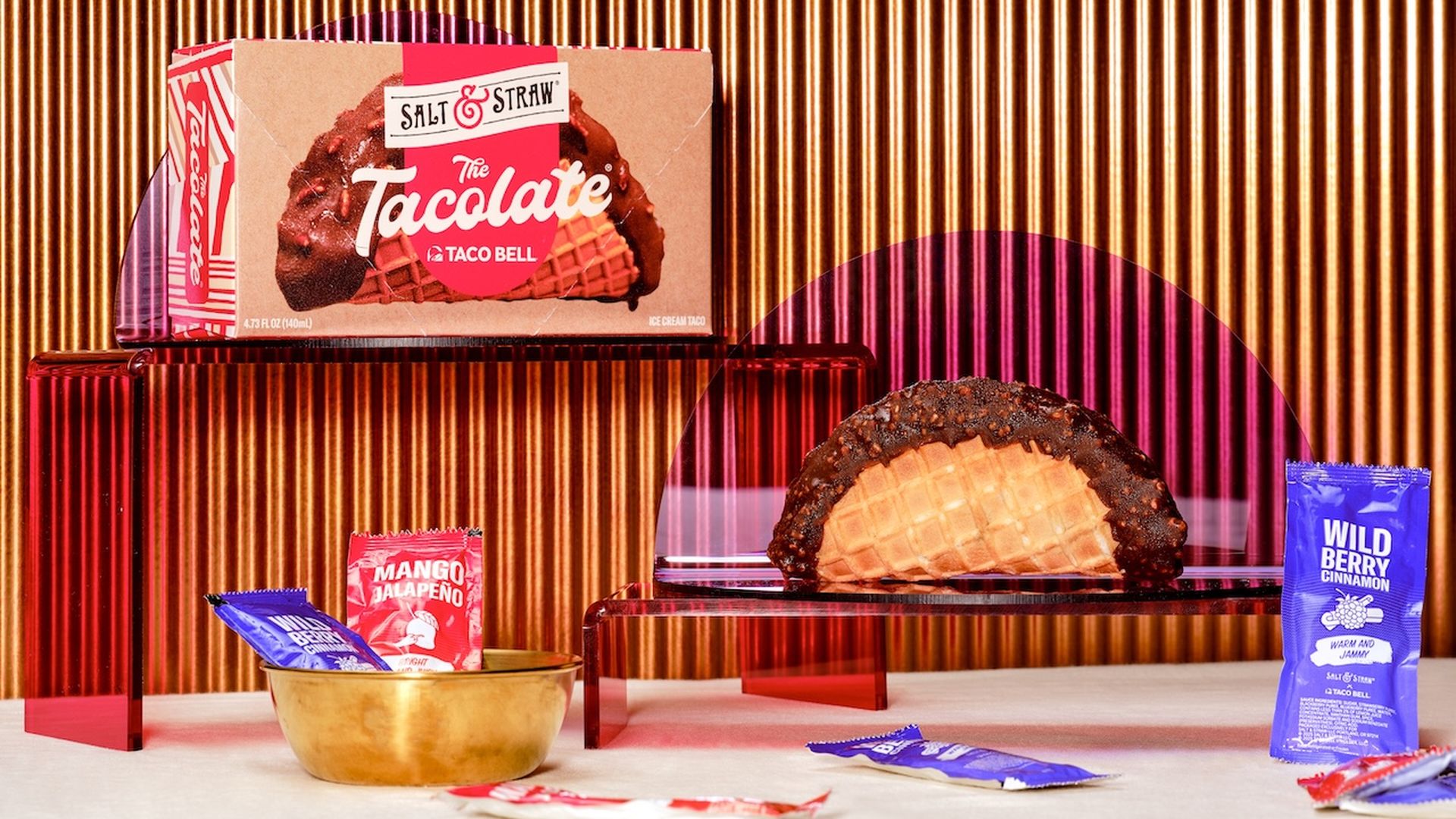 Salt & Straw x Taco Bell Tacolate ice cream taco in a waffle shell dipped in chocolate, with Mango Jalapeño and Wild Berry Cinnamon sauce packets on a red and gold ribbed background.