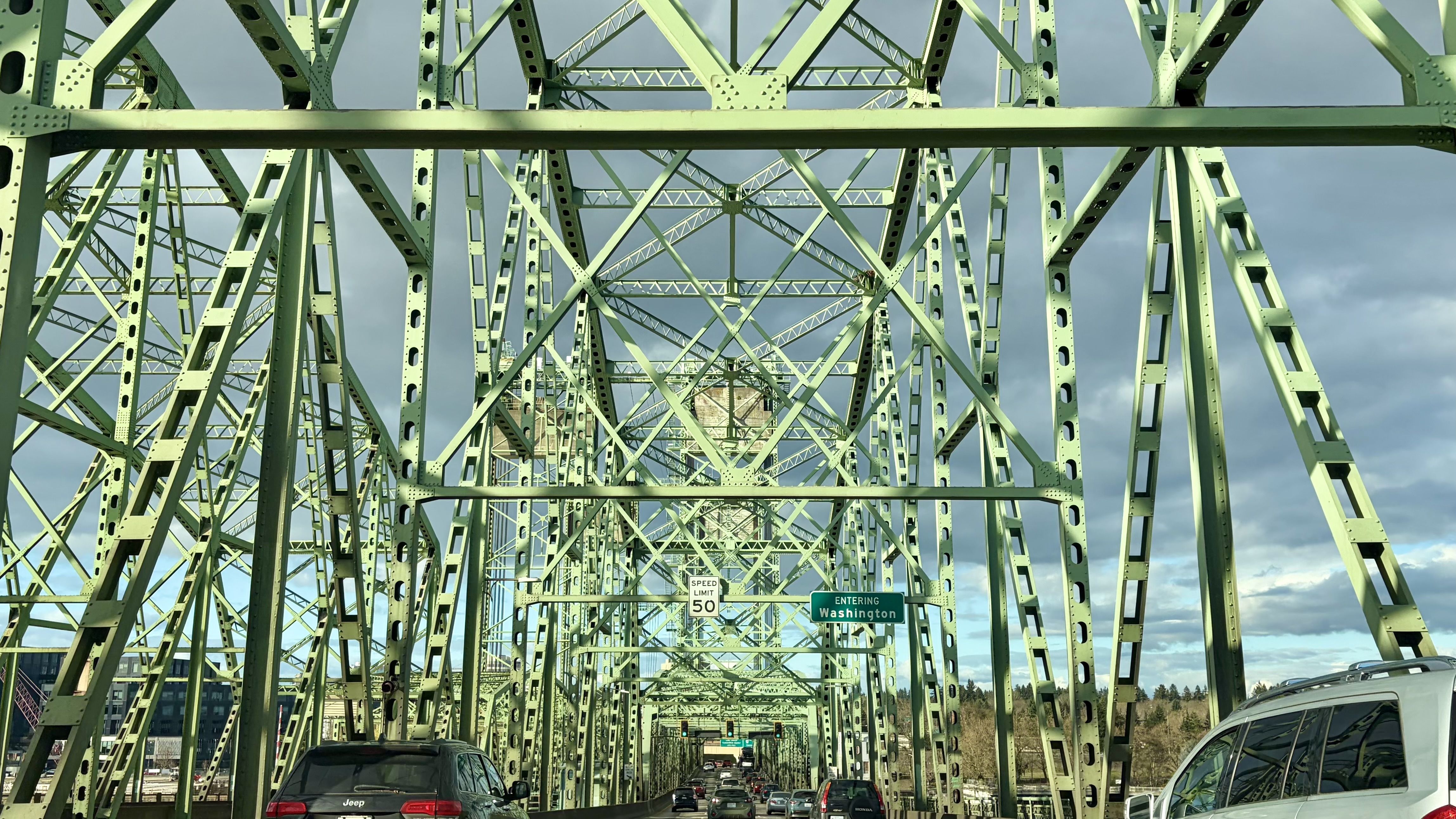 View along a green steel truss bridge crowded with cars; crisscrossed beams create a lattice under a gray, cloudy sky. Sign reads "Entering Washington" and a 50 mph speed limit.