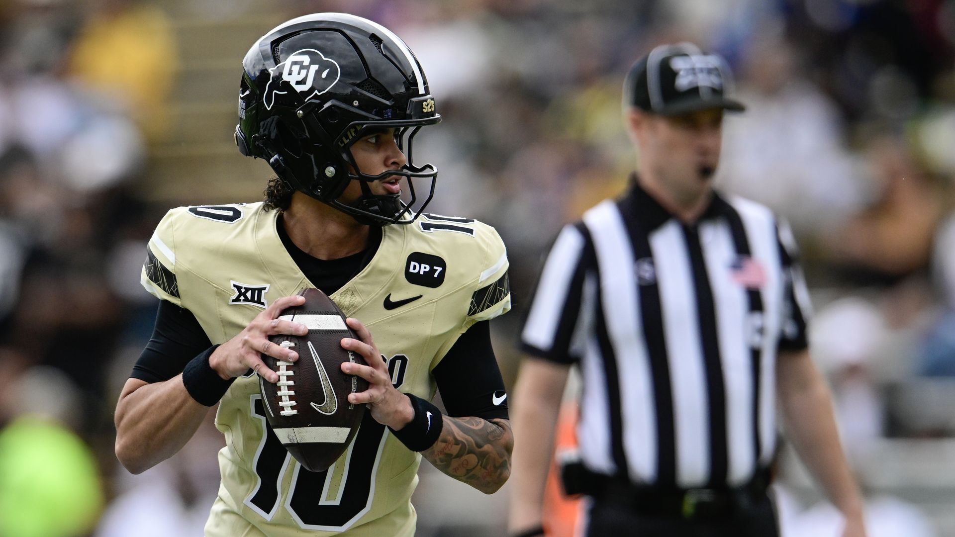 A college football player in a gold uniform and black helmet with a CU logo grips a football, looking to the left, as a striped referee stands blurred in the background.