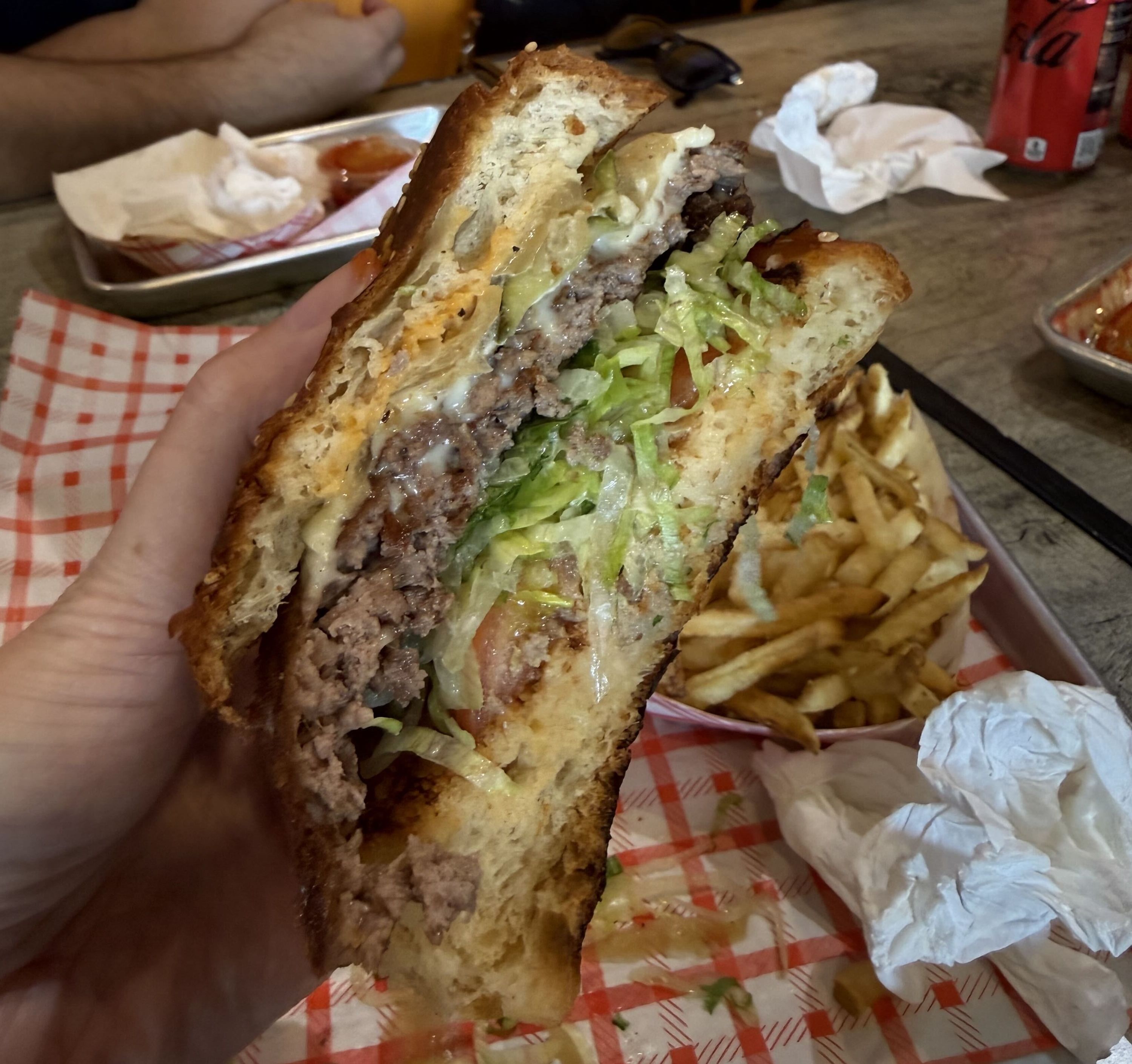 A hand holds a thick burger with lettuce, tomato, melted cheese and beef on a toasted bun; fries and napkins visible in the background.