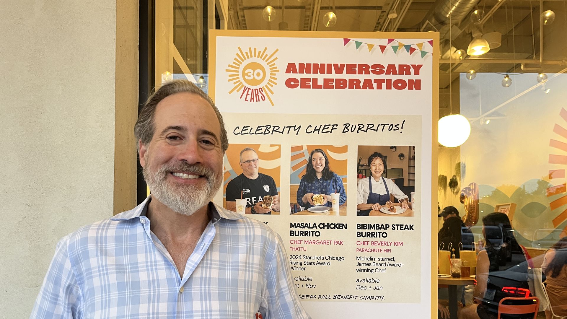 Smiling man with gray hair and beard in a light blue plaid shirt stands next to a colorful 30th anniversary celebration sign for Burrito Beach featuring celebrity chef burritos.