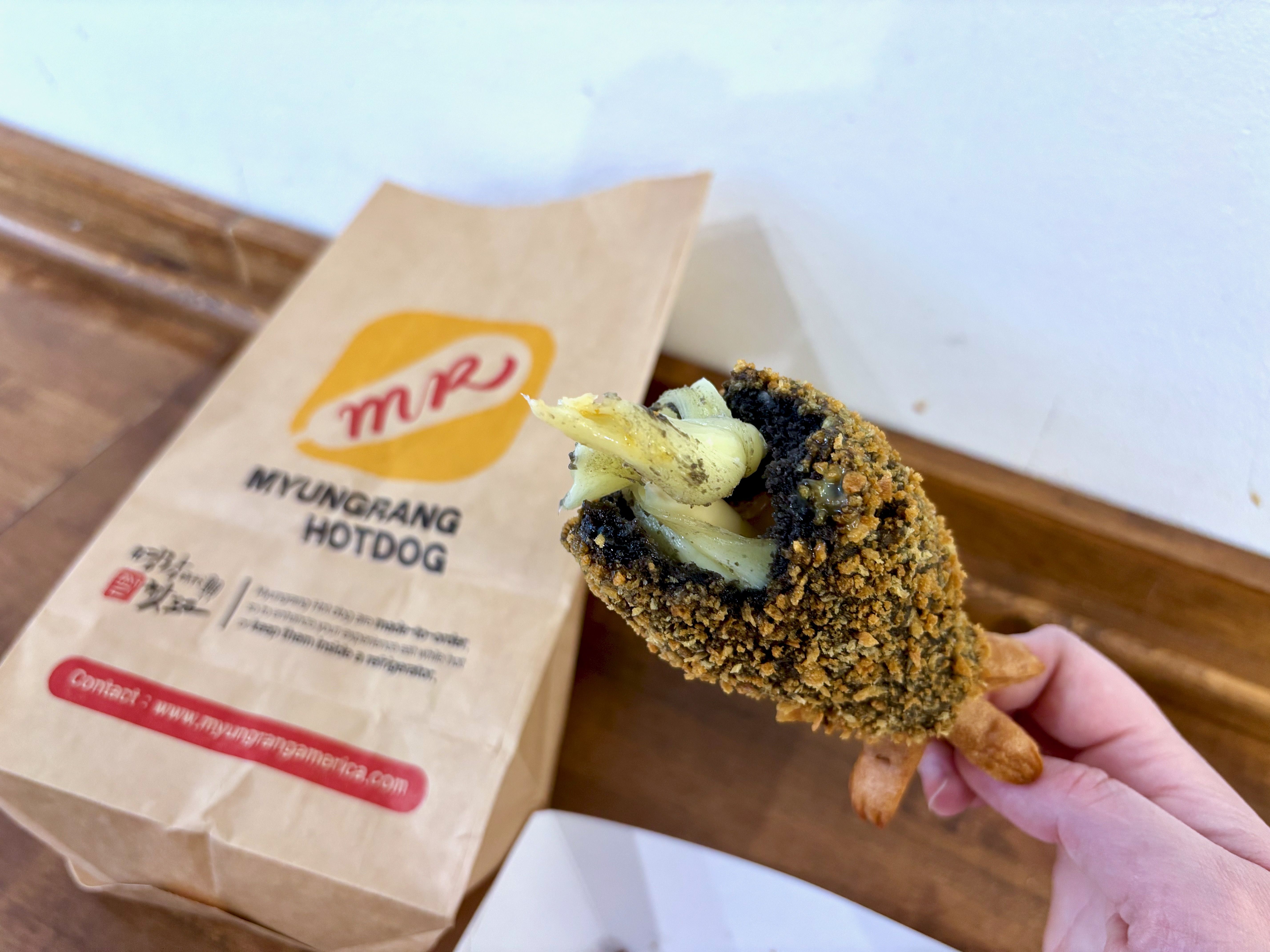 Hand holding a fried, breaded hot dog with dark breading and melted cheese inside, in front of a brown paper bag labeled "Myungrang Hotdog" on a wooden surface.
