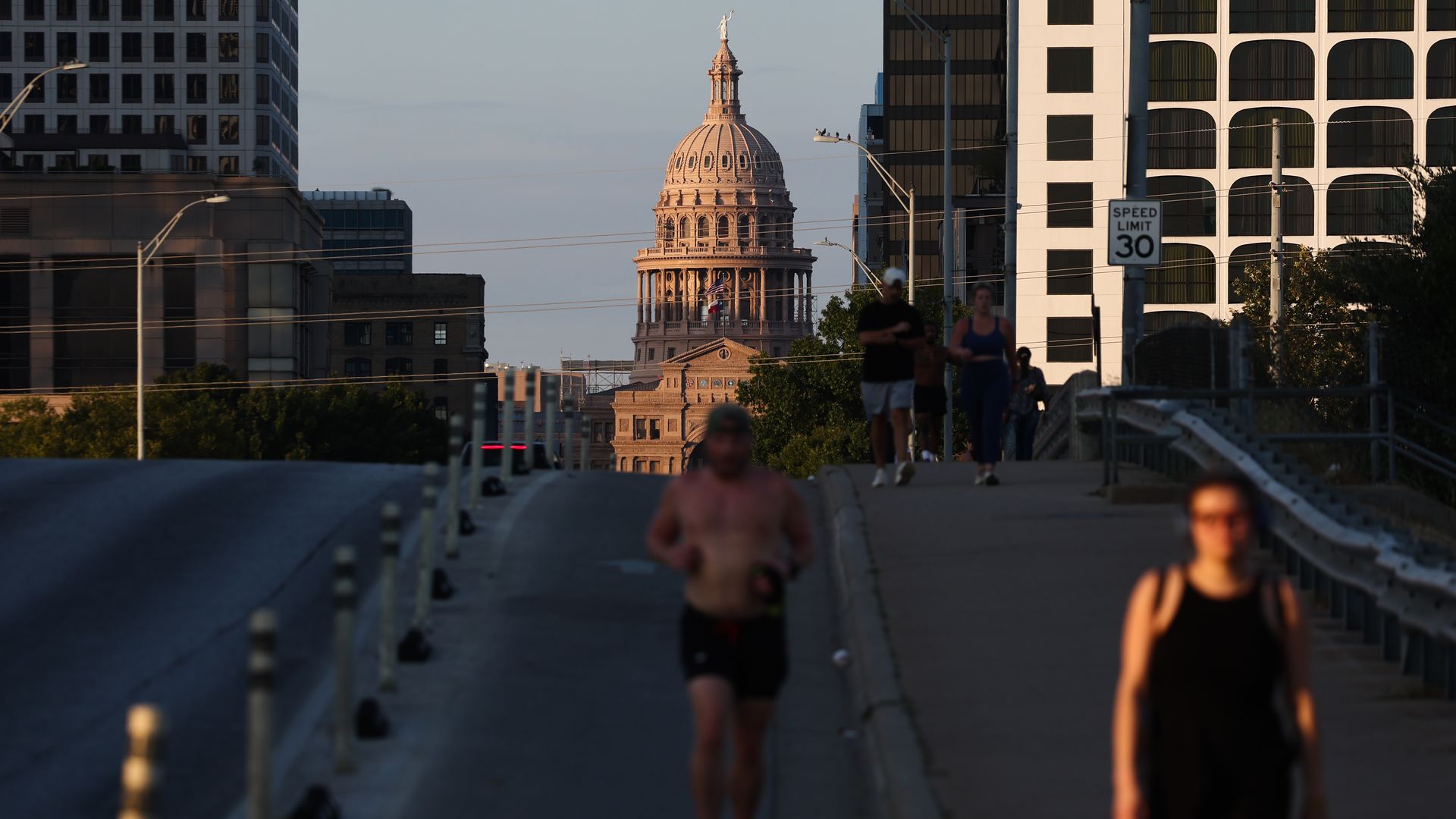 People jogging and walking on a road and sidewalk in a city with the Texas State Capitol building lit by sunset in the background, surrounded by skyscrapers and a speed limit 30 sign