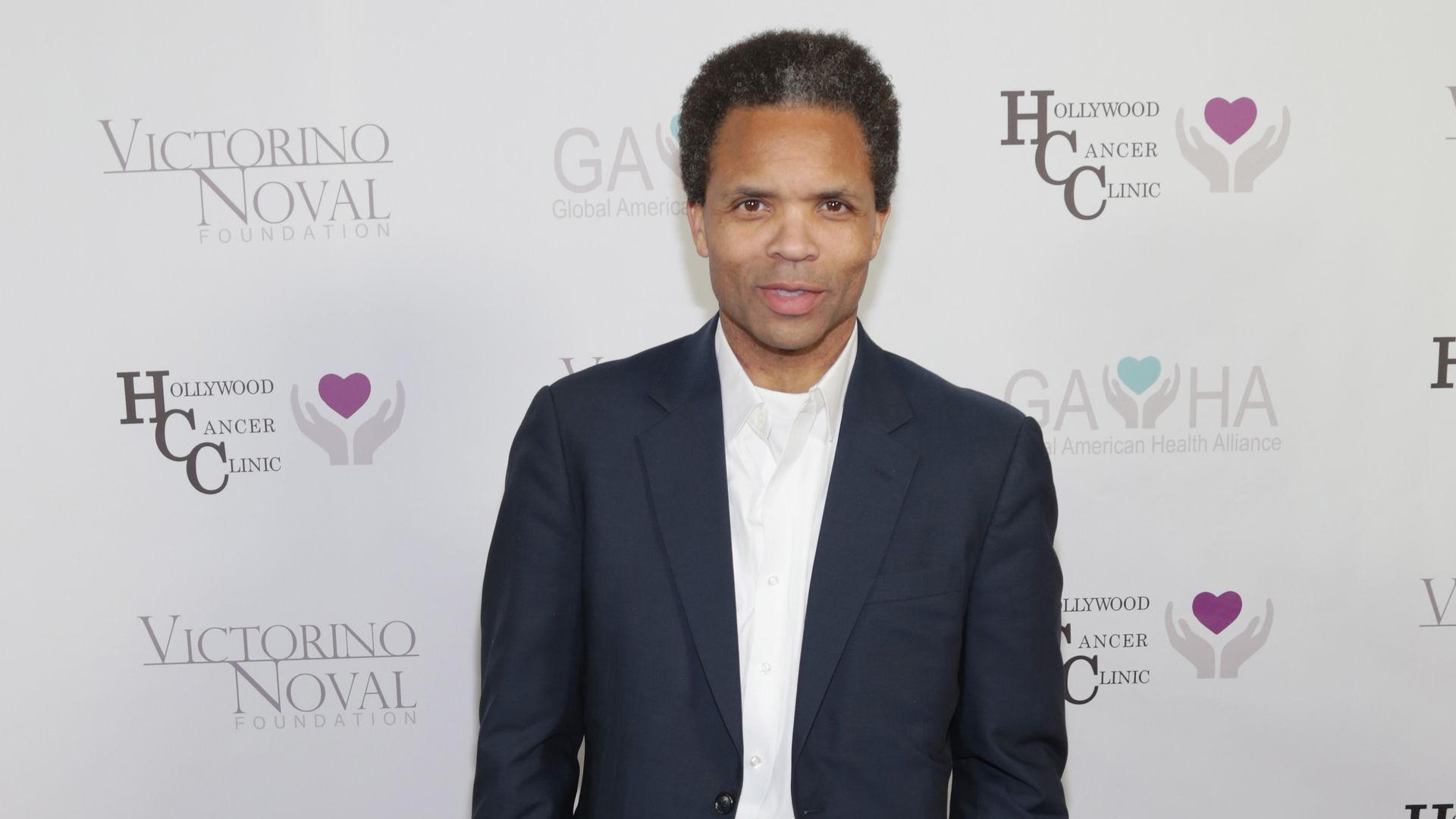 Jesse Jackson Jr., wearing a blue suit with no tie, stands in front of a white backdrop with logos on it.