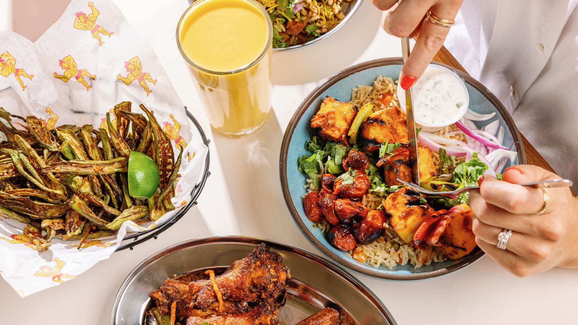 Top-down view of a vibrant Indian meal: bowls of spiced chicken with rice and cilantro, a side of fried okra with lime, a creamy dipping sauce, and a glass of mango lassi; a hand serves with a spoon.