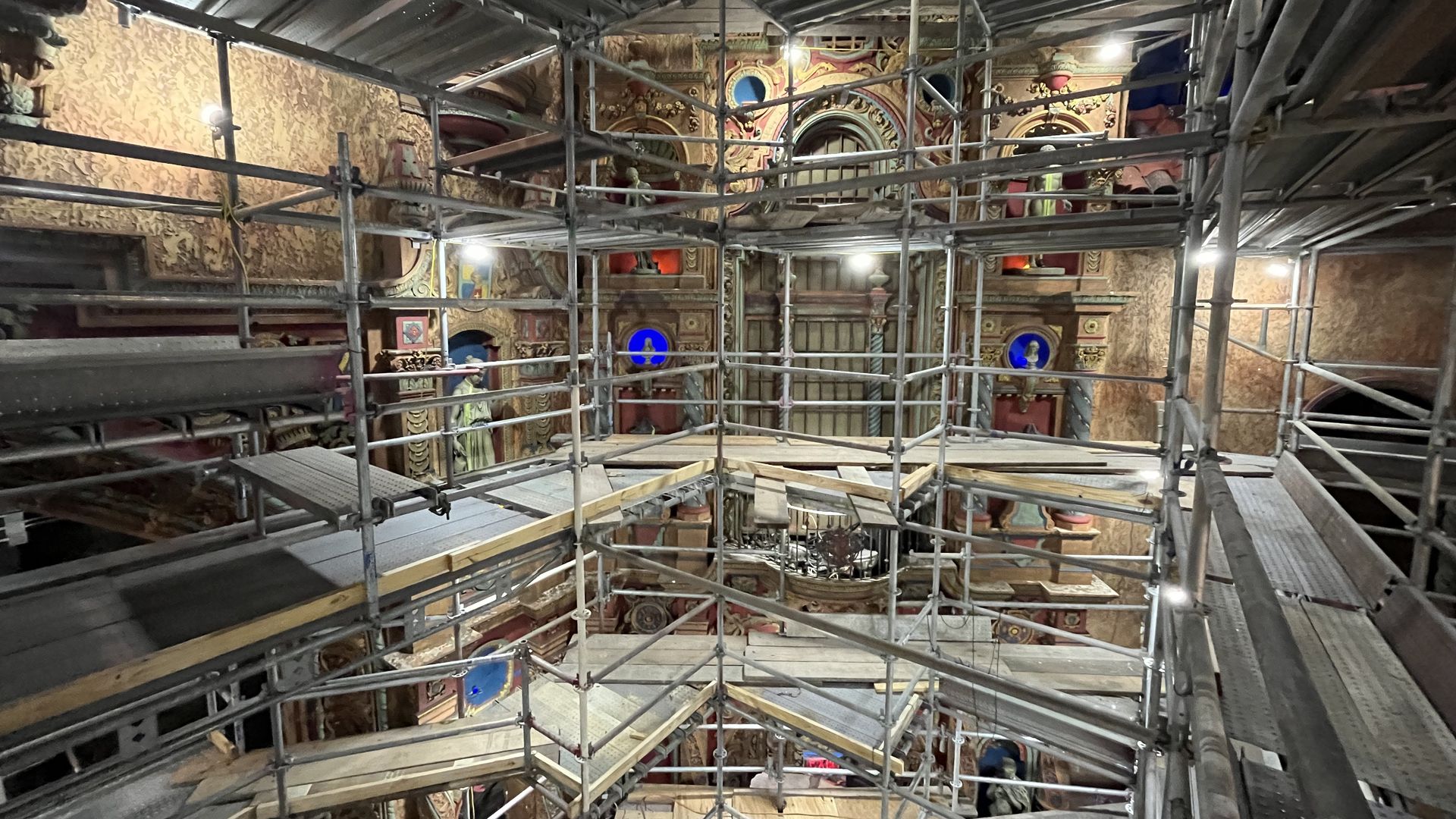Interior of a historic space under dense metal scaffolding; ornate gold arches and murals are visible behind the lattice, warm lights, blue circular medallions, and wooden planks on the floor.