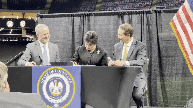 Three officials in suits sitting at a table with a blue State of Louisiana banner, signing documents, with a black curtain backdrop and a U.S. flag visible on the right.