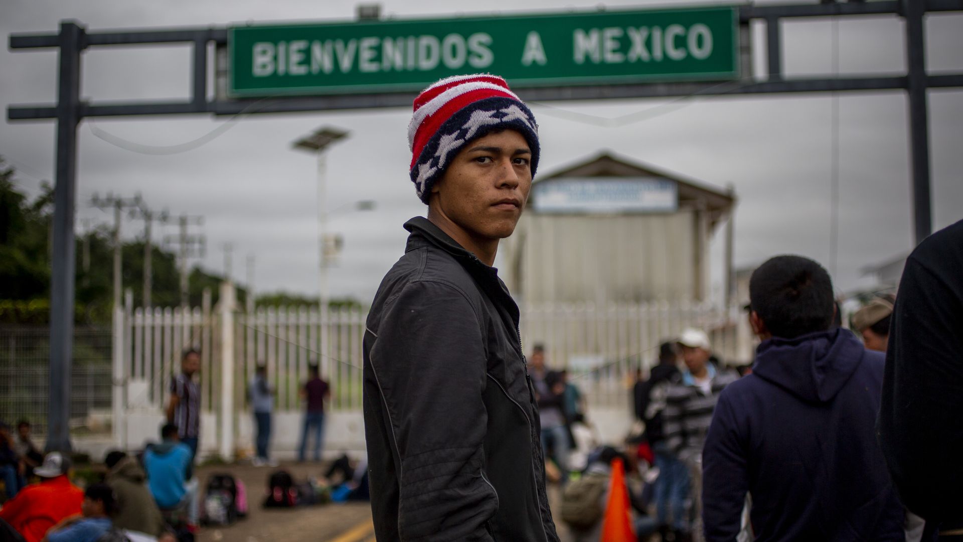 A teenage migrant stands on a bridge with a blurred sign in the background which says "welcome to Mexico" in spanish.