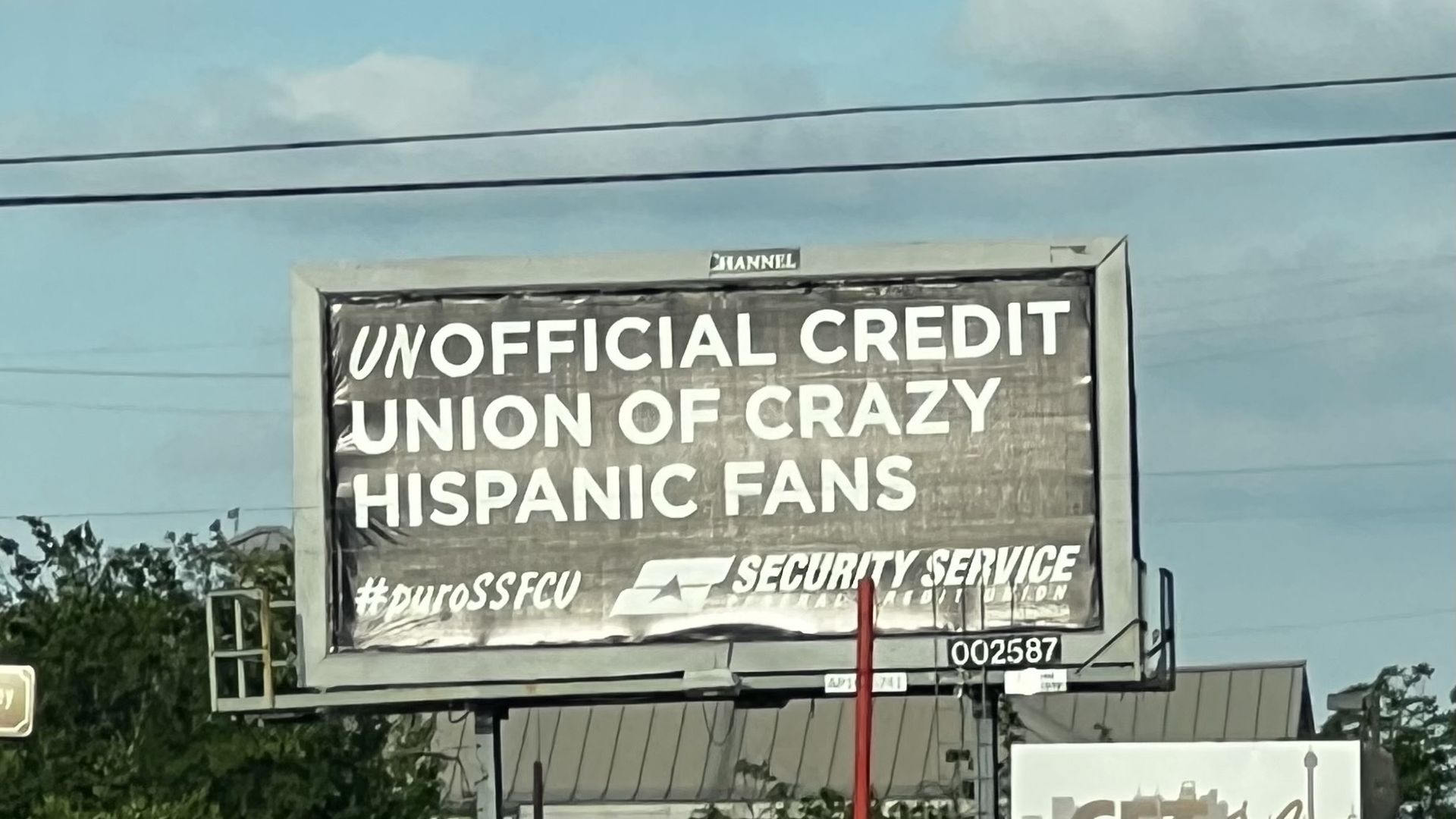 Outdoor billboard with bold white text on a dark background reading "UNOFFICIAL CREDIT UNION OF CRAZY HISPANIC FANS," framed in gray under a blue sky with power lines.