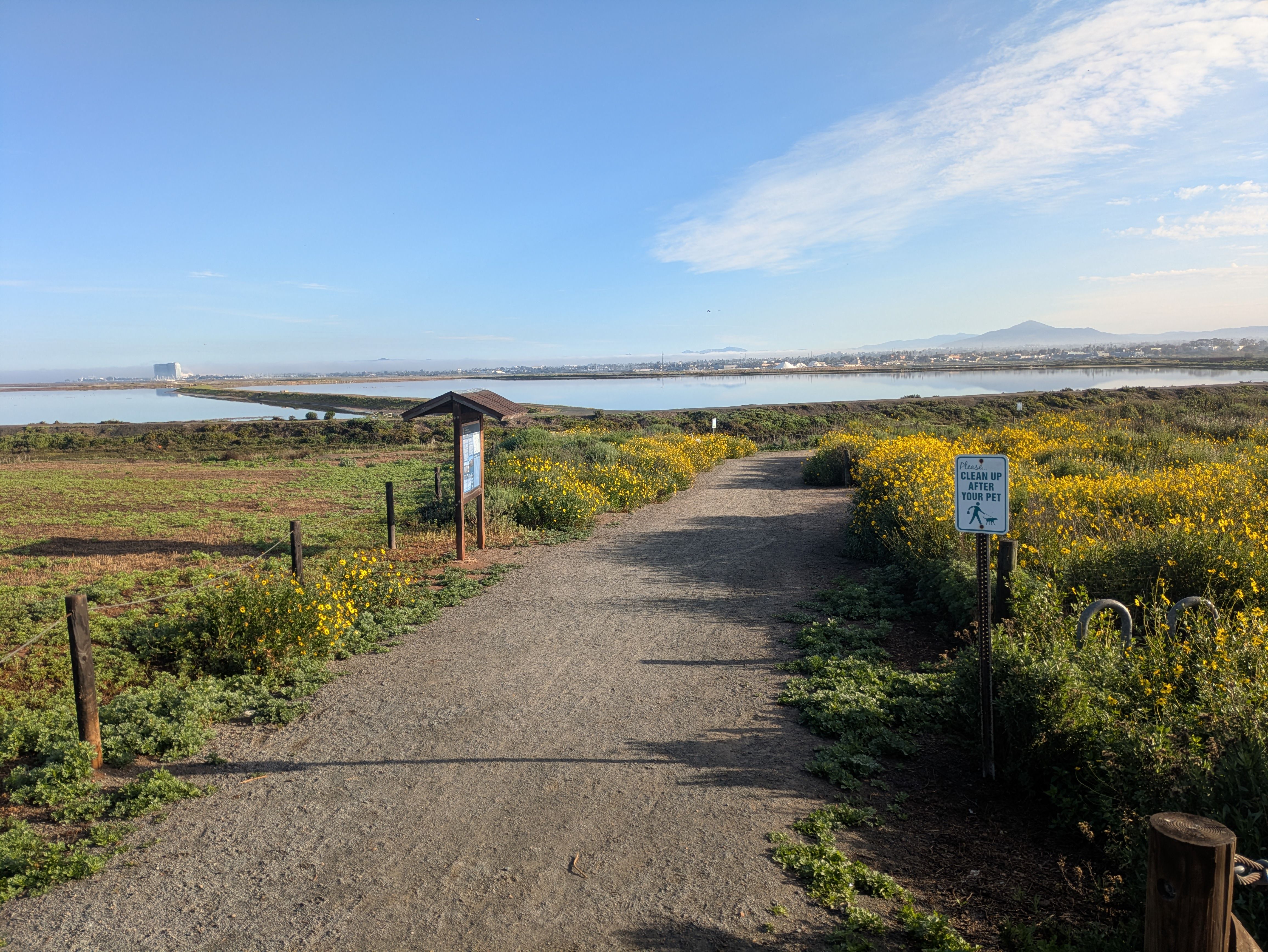 A dirt path through yellow wildflowers leads to a lake under a blue sky; a sign reads "Please clean up after your pet" on the right, with hills and a distant building in the background.