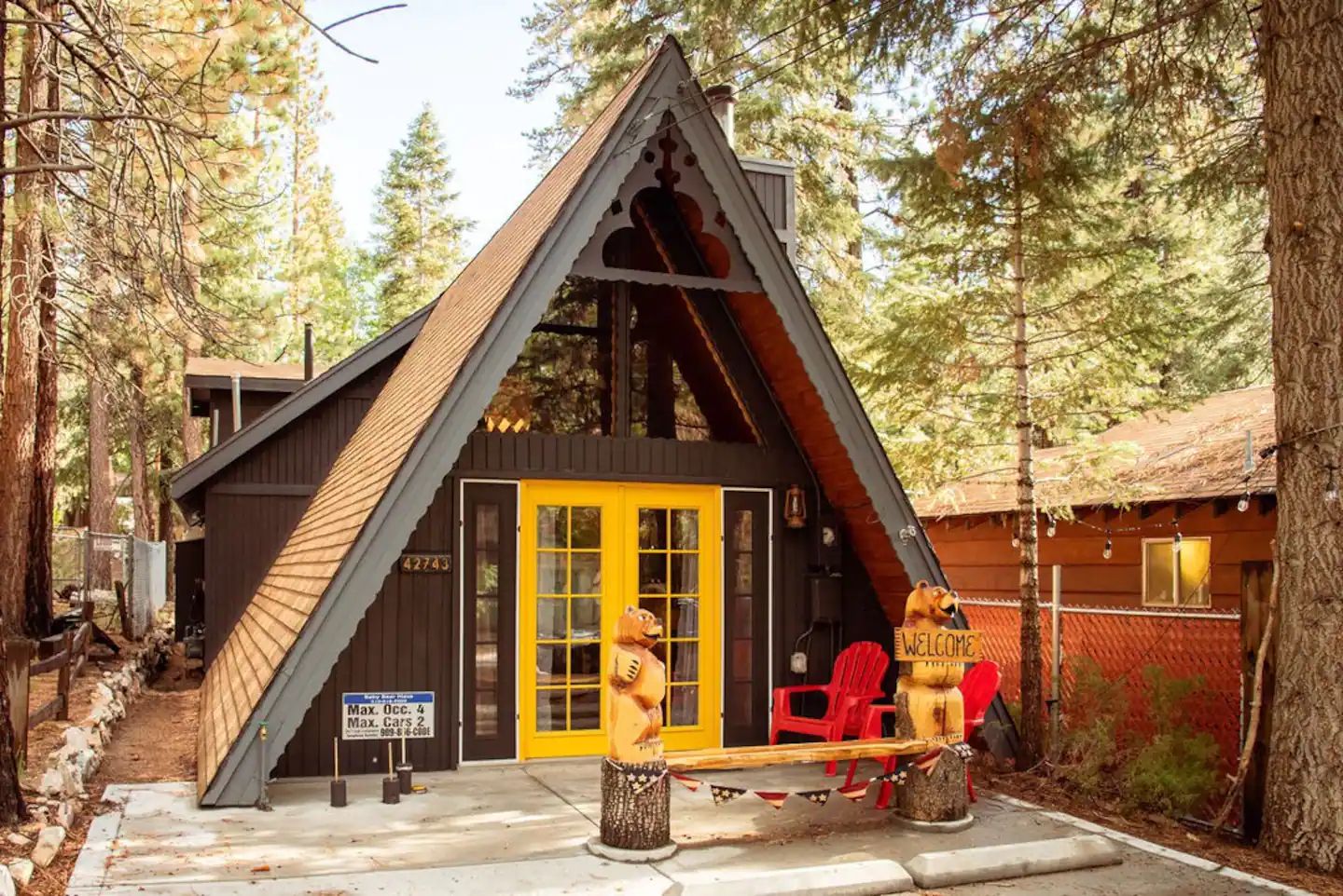 Two wooden carved bears welcome guests to an A-frame cabin in the woods with bright yellow doors.