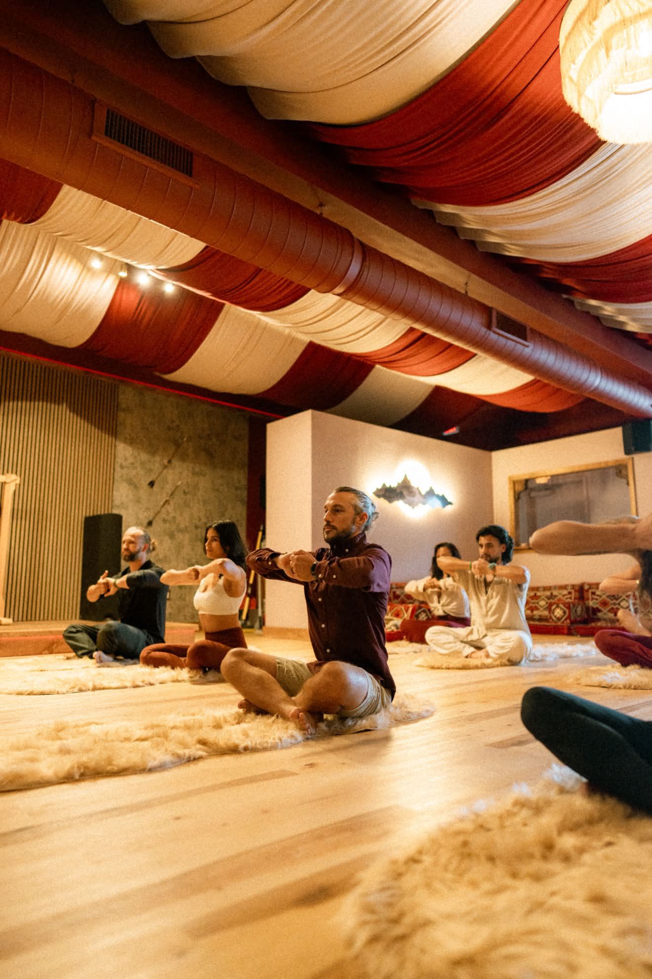 Group of people practicing seated yoga in a studio, with a red and cream fabric-draped ceiling, warm lighting, and a wooden floor.