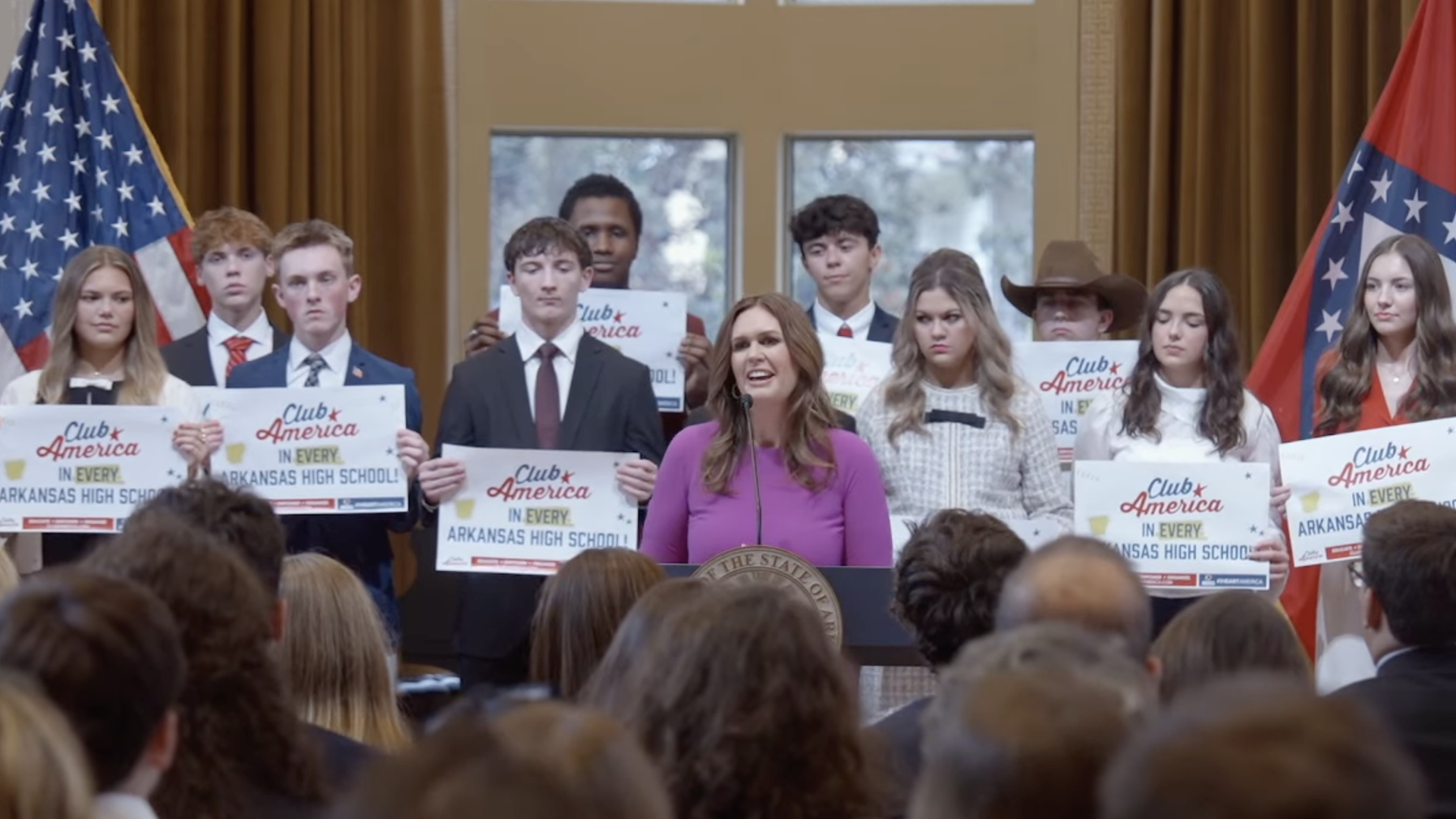 Gov. Sarah Huckabee Sanders speaks at a podium flanked by U.S. and Arkansas flags, with students behind her holding "Club America in Every Arkansas High School" signs.