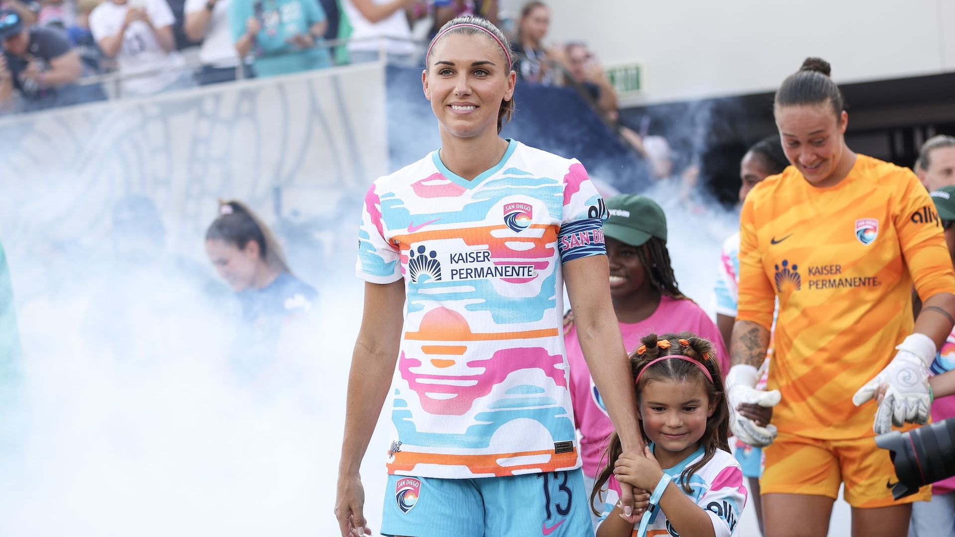 Alex Morgan and her daughter walk out of the tunnel onto the field with smoke and Wave players behind her.