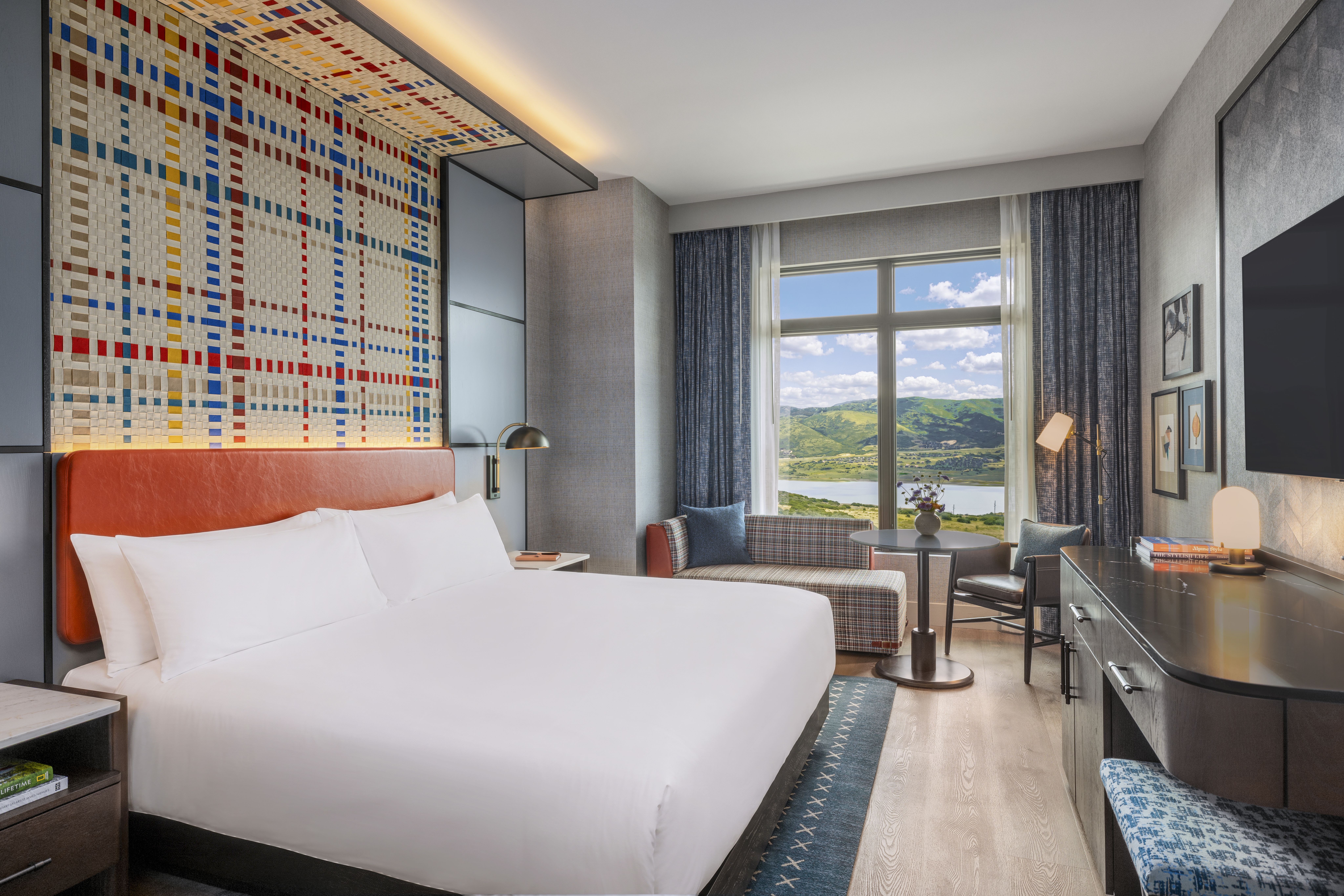 Modern hotel room with a white bed and orange headboard, a large colorful mosaic wall, gray walls, a window with blue curtains showing green hills, and a small seating area.