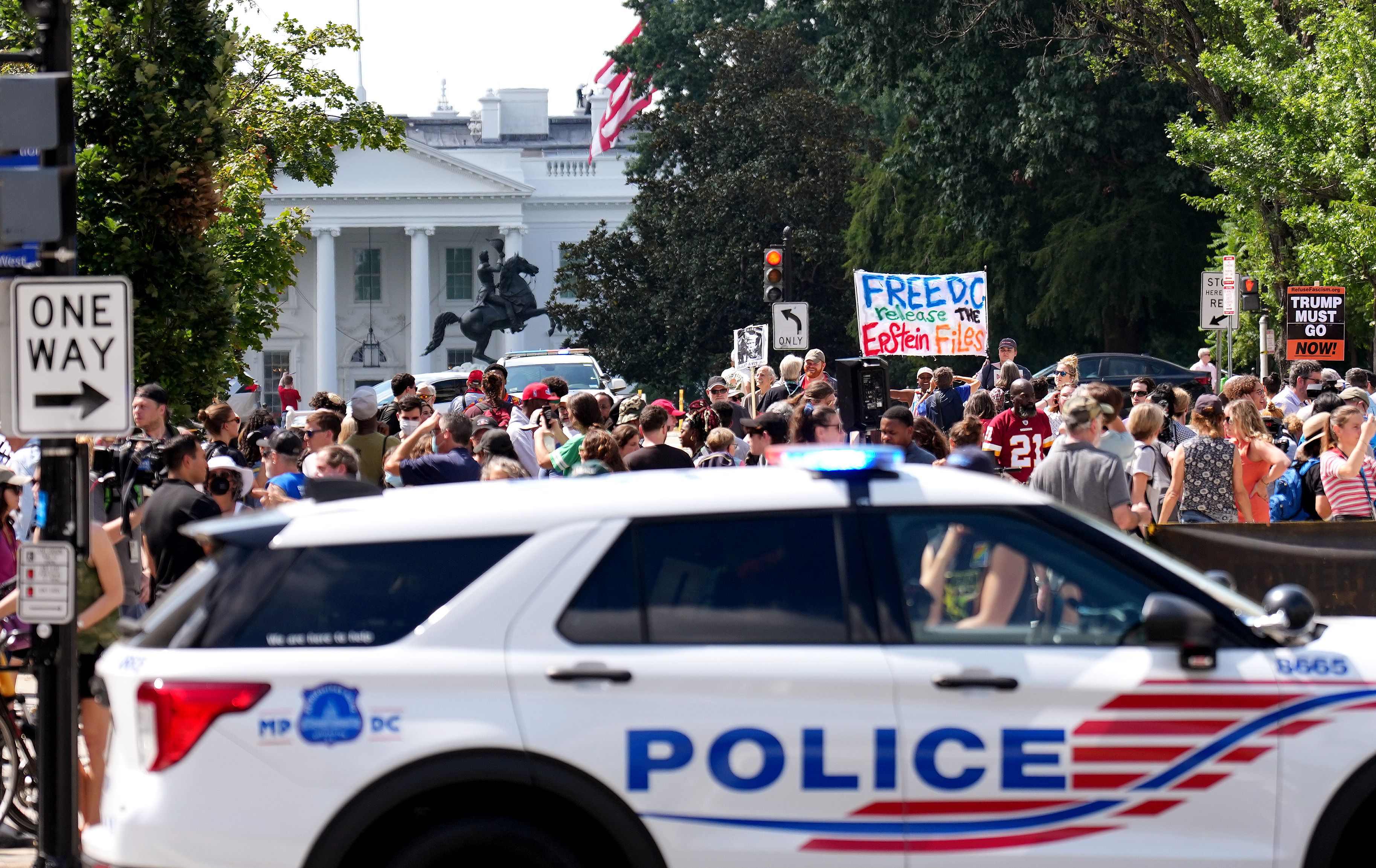 Crowd of people with signs including "FREE DC release the Epstein Files" and "TRUMP MUST GO NOW!" near the White House, with a police car in the foreground and a one way sign.