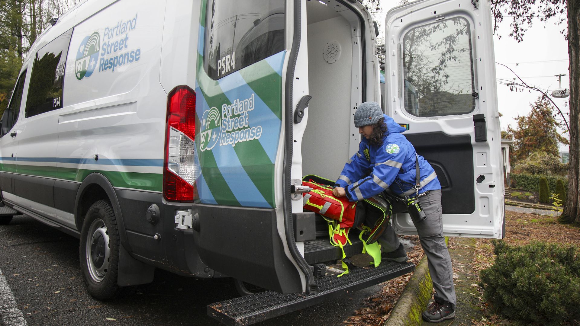 Person in blue jacket and gray hat loading red and yellow emergency gear into the back of a white van labeled "Portland Street Response" with green and blue stripes.