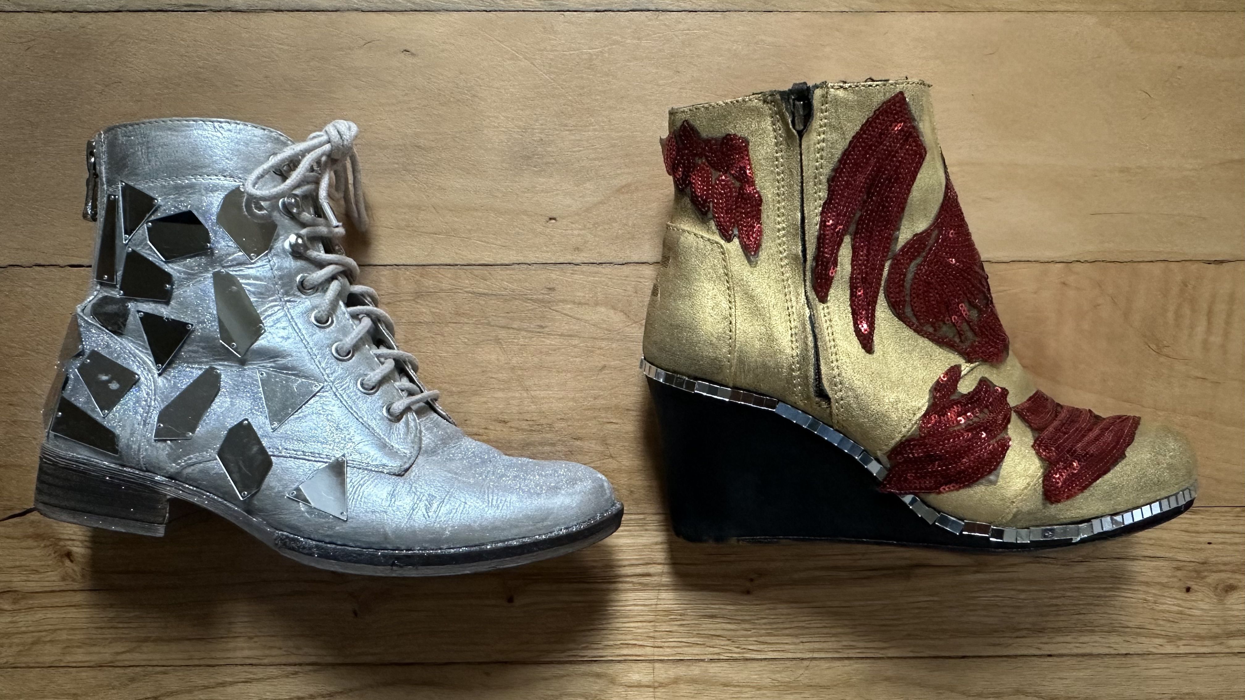 A silver lace-up boot embellished with mirrored pieces lays on the ground next to a gold-painted boot covered in sequin patches shaped like red feathers.