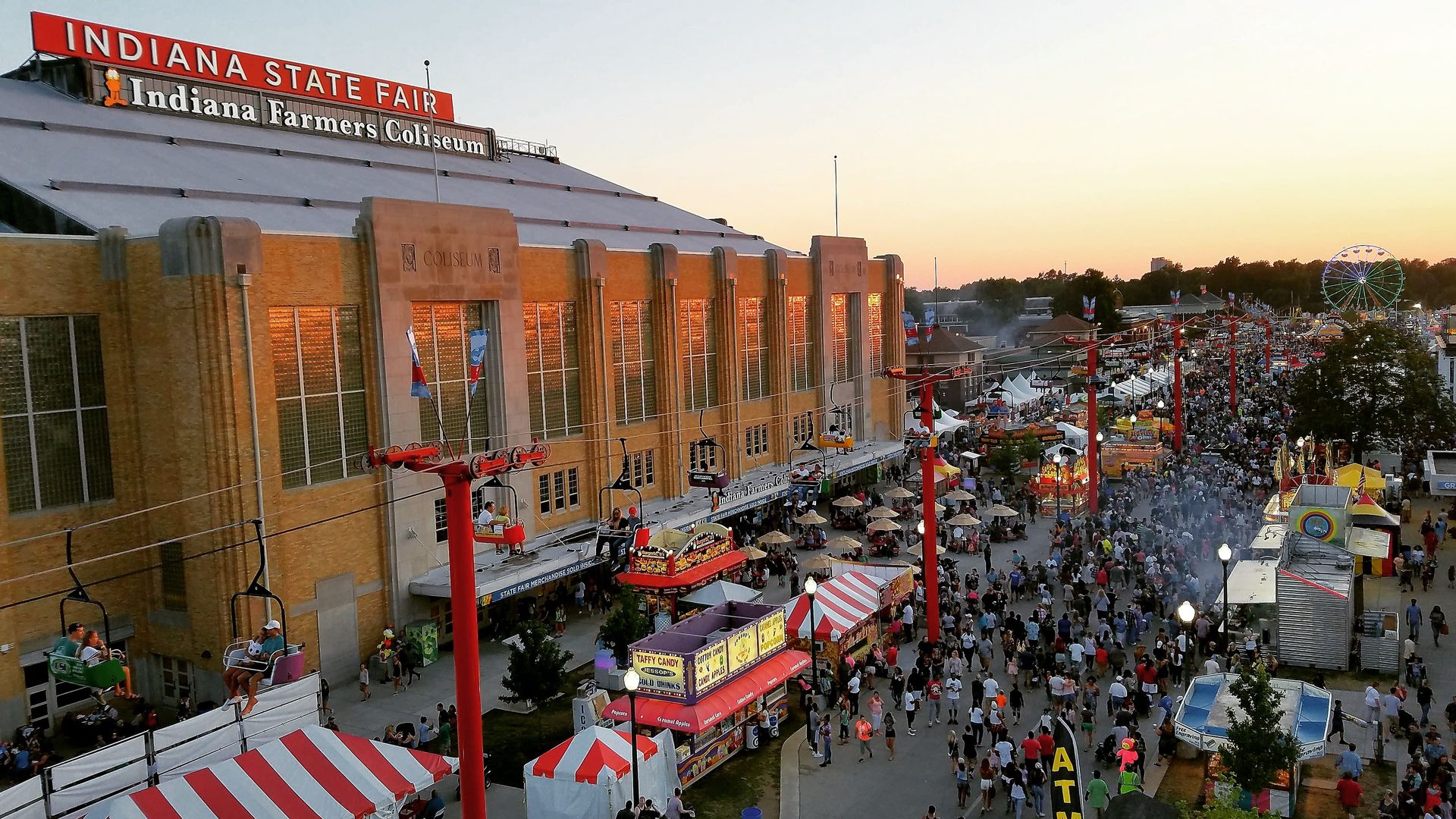 Hundreds of people walk main street at the Indiana State Fair at dusk, with a ferris wheel in the background.