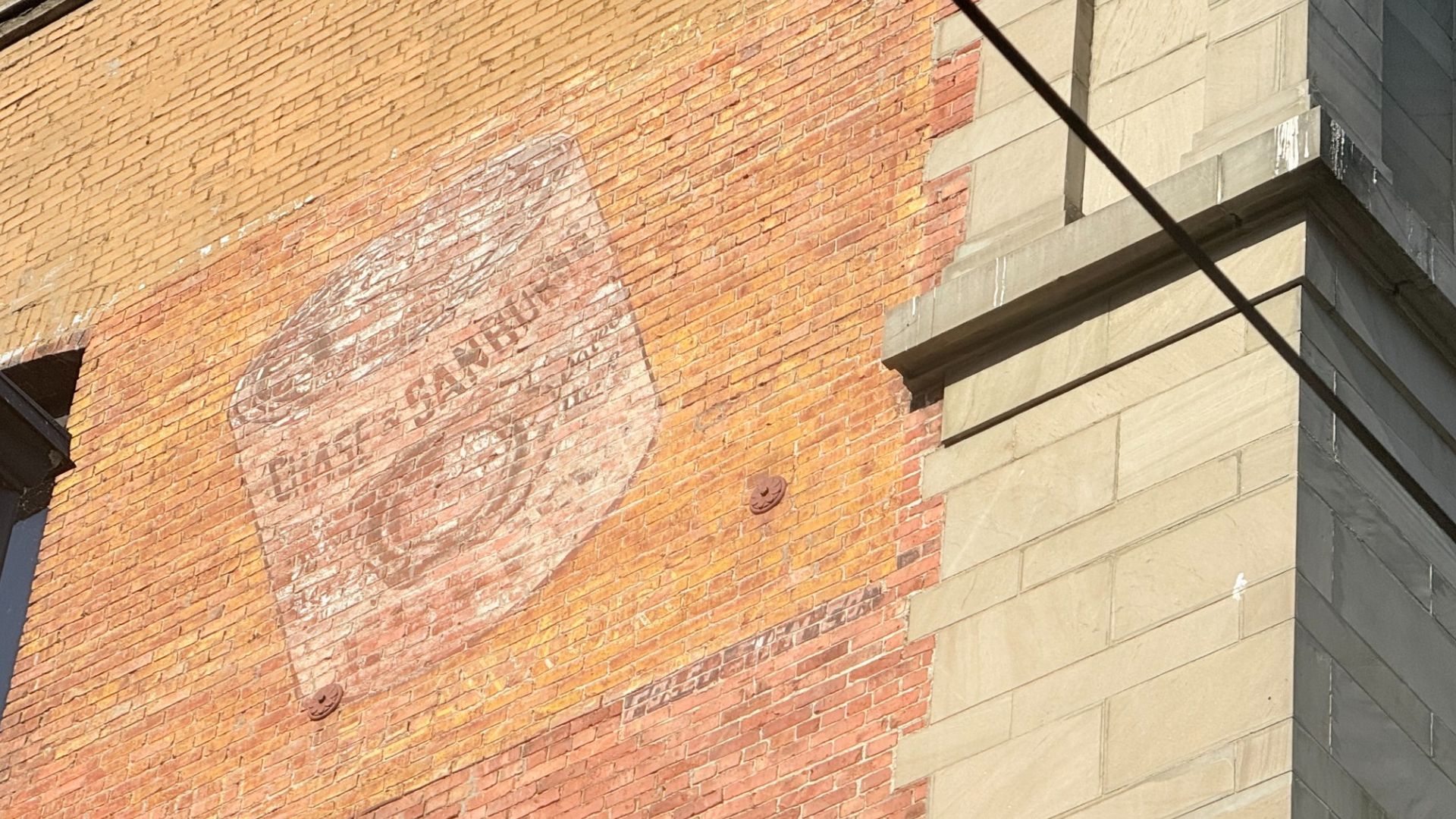 Orange brick wall with a large faded circular mural or logo. Beige stone building corner on the right; dark diagonal wires cross the scene.