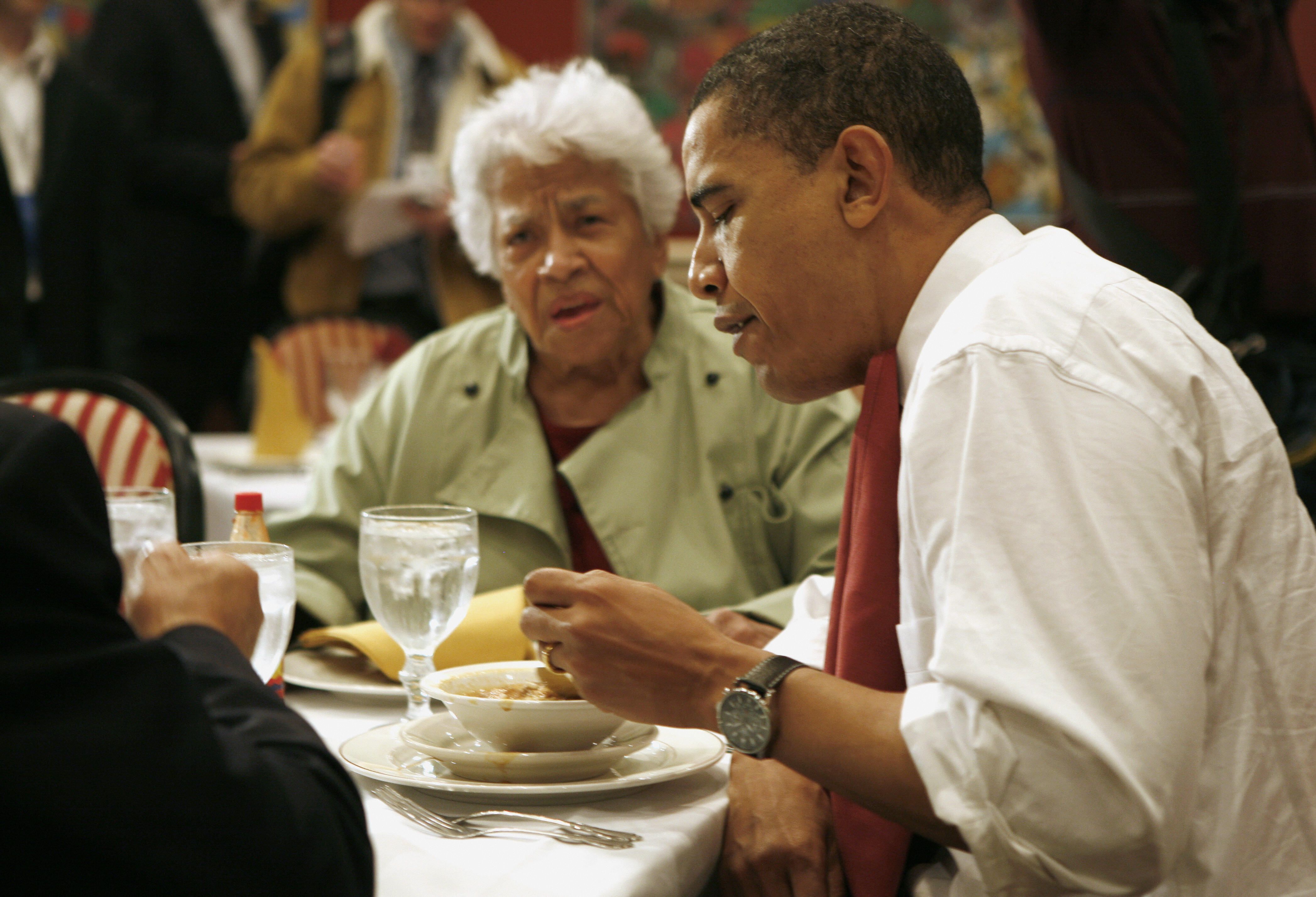 Barack Obama in white shirt and red tie eating soup at a restaurant table with Leah Chase wearing a light green coat seated nearby, several water glasses on the table.