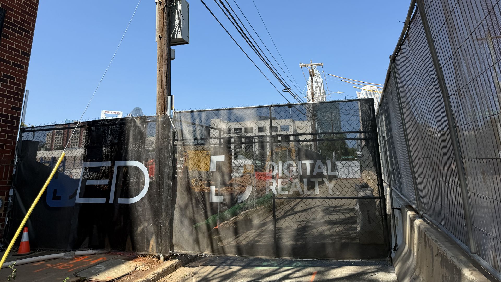 Urban alley with a tall wooden utility pole and overhead power lines, brick wall on the left, corrugated fence on the right, and distant skyscrapers beneath a clear blue sky.