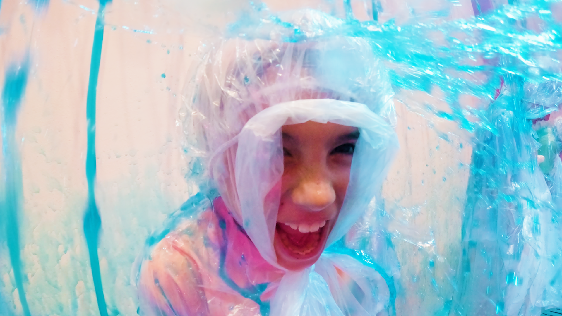 Child joyfully laughing inside a large clear inflatable ball filled with blue liquid, wearing a transparent plastic rain poncho and hood indoors on a blue and pink background.