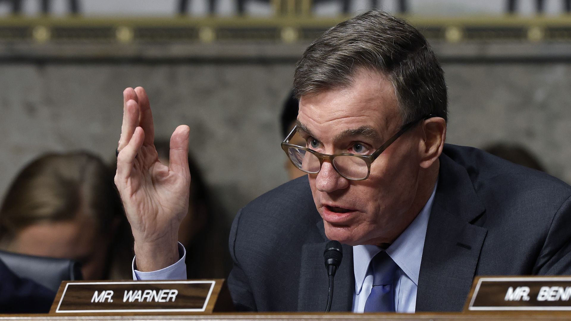 Sen. Mark Warner, wearing a navy jacket, a light blue and white checked shirt and navy tie, gestures with his right hand at a brown, wooden desk in front of a sign reading "MR. WARNER."