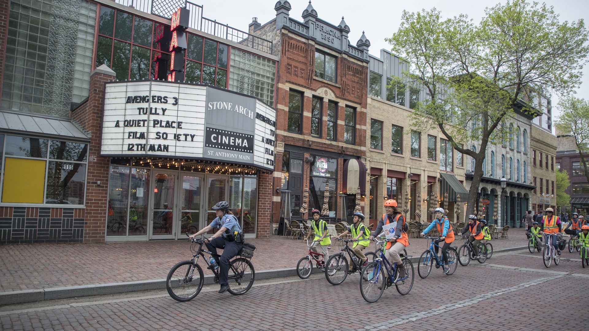 About 10 people on bicycles wearing bright yellow and orange vests bike down Main Street, with bricks on the street and historic buildings in the background. A movie theater marquee shows showtimes. 