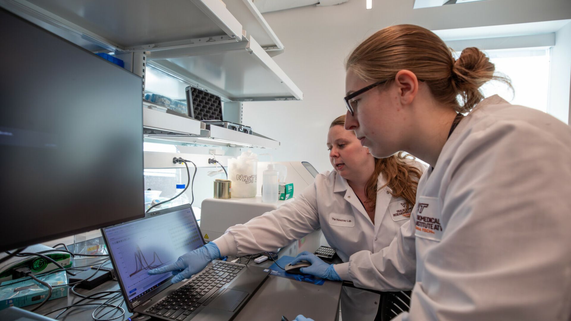 Two scientists in white lab coats and gloves work at a bench with laptops and graphs; shelves of equipment behind them in a bright, modern laboratory.
