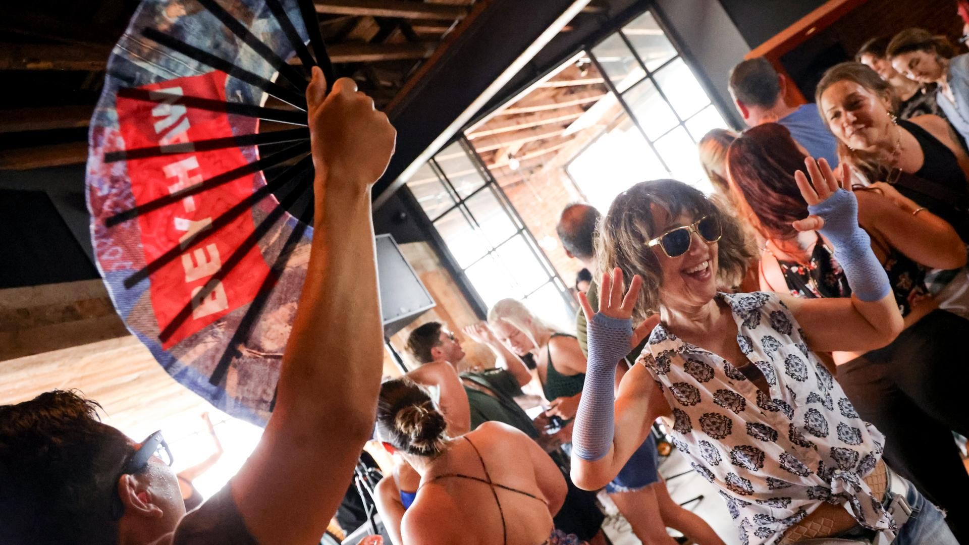 Dancers with a fan and sequined fannypack are part of a group moving in front of bright windows at a coffee rave in Los Angeles