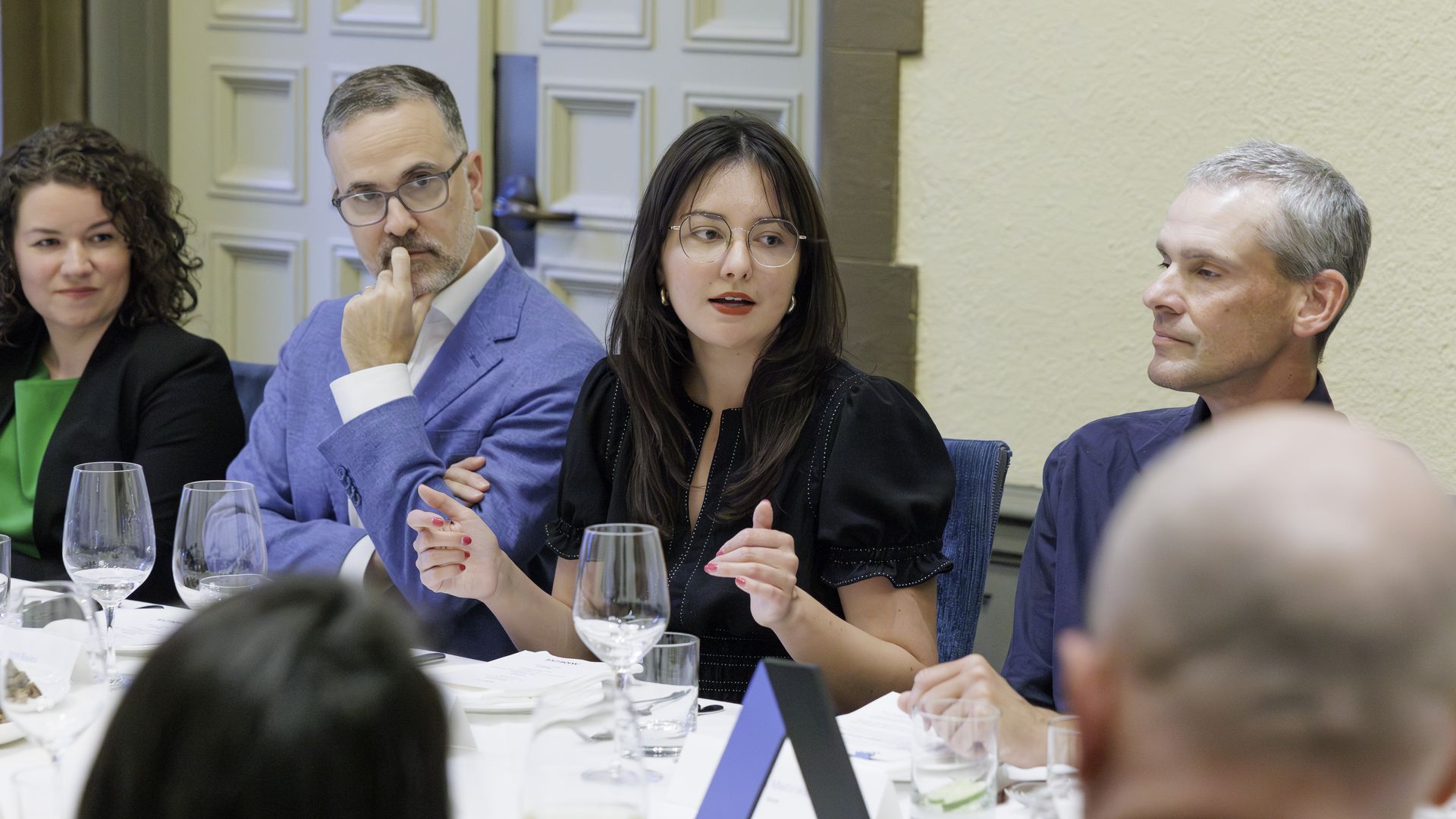 Four people sit at a formal round-table; the center woman with glasses in a black blouse speaks, flanked by a man in a blue blazer and another man, with a woman in green on the left.