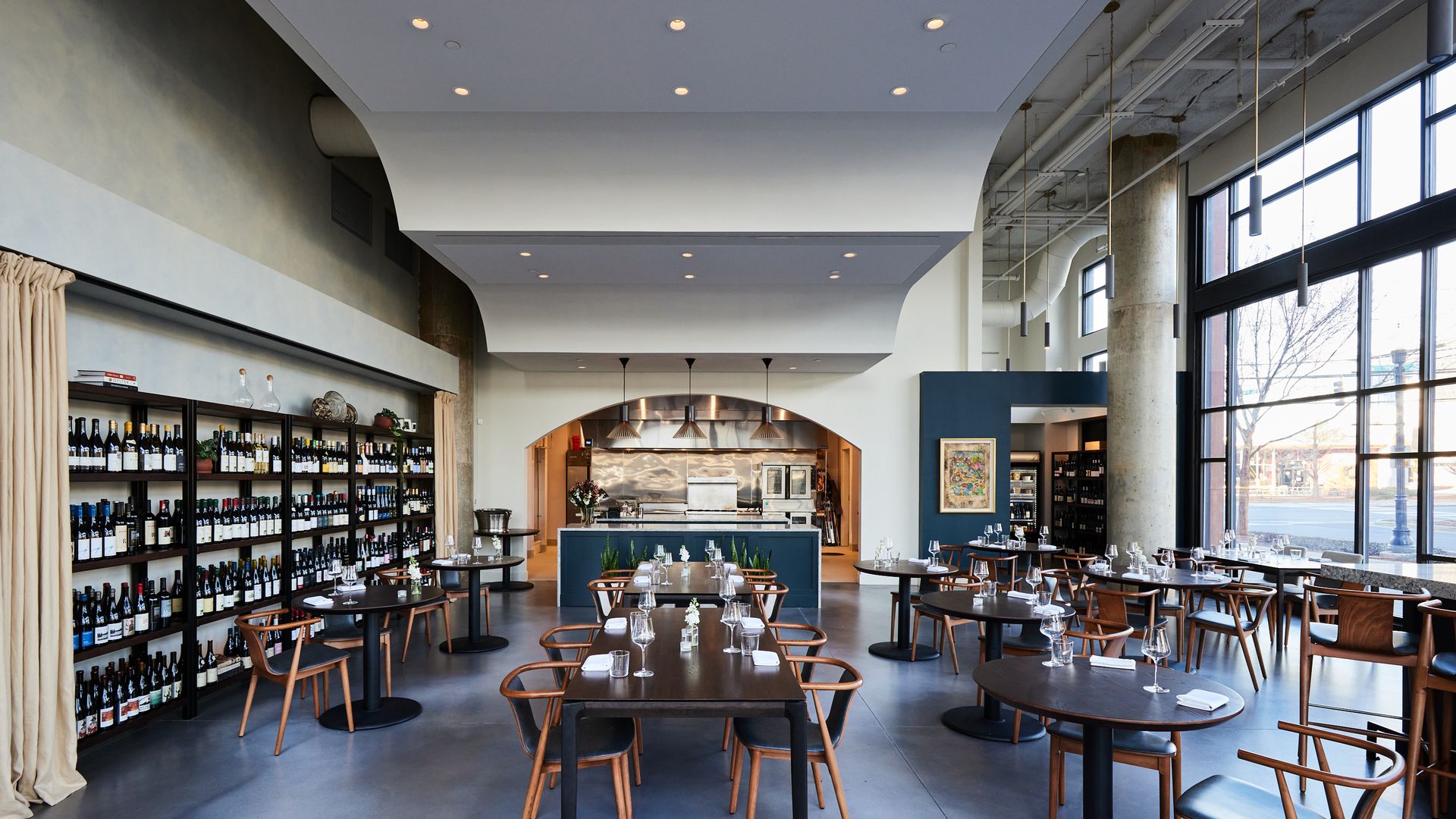 Modern restaurant interior with wooden chairs and dark tables set with glasses and napkins, large windows, wine shelves on the left, and open kitchen in back with pendant lights.