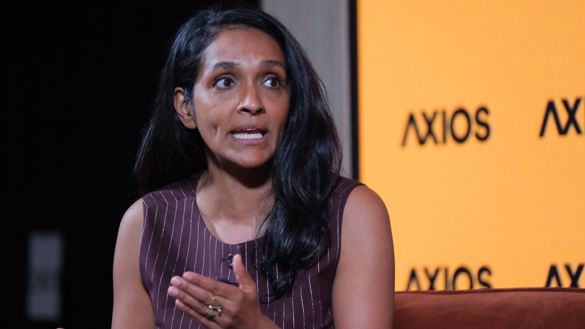 Mayoral candidate Nithya Roman speaking on stage, hands gesturing. She wears a maroon striped sleeveless dress; a bright orange Axios backdrop is visible on the right.