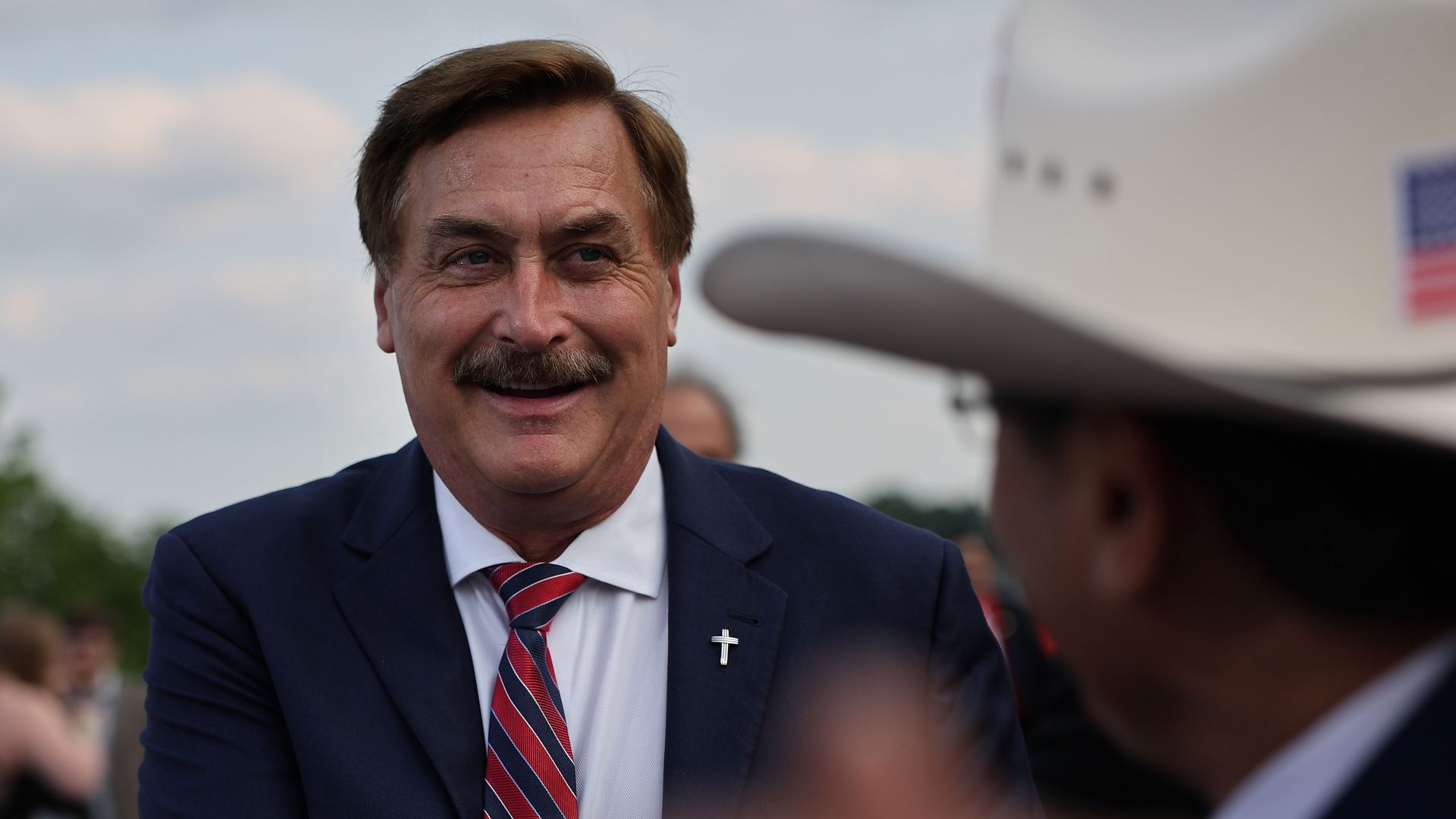 Mike Lindell, wearing a dark suit and red and blue tie with a cross pin, smiles. In the foreground, a man wears a cowboy hat. 