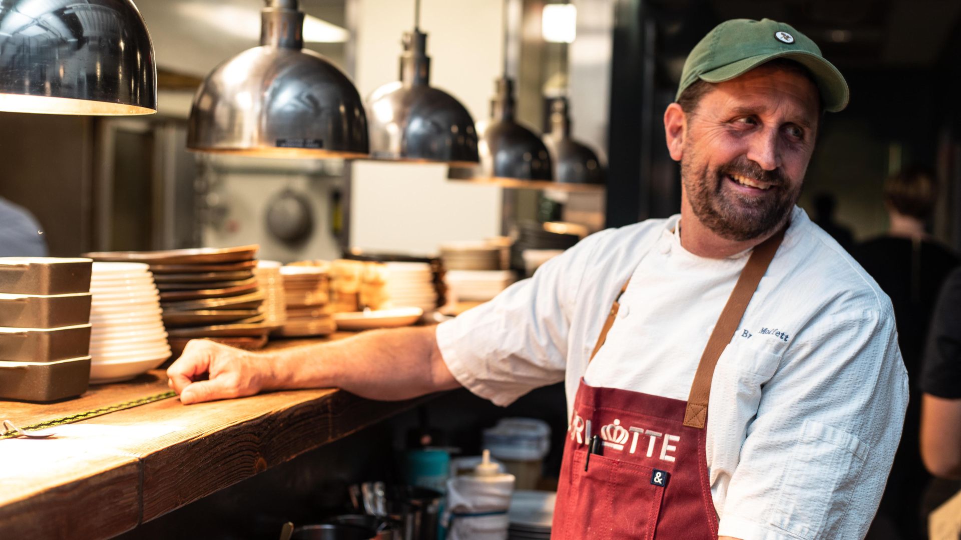 A man in a green cap and red apron leans at a wooden counter in a busy kitchen, with stacked plates and shiny hanging heat lamps behind him.