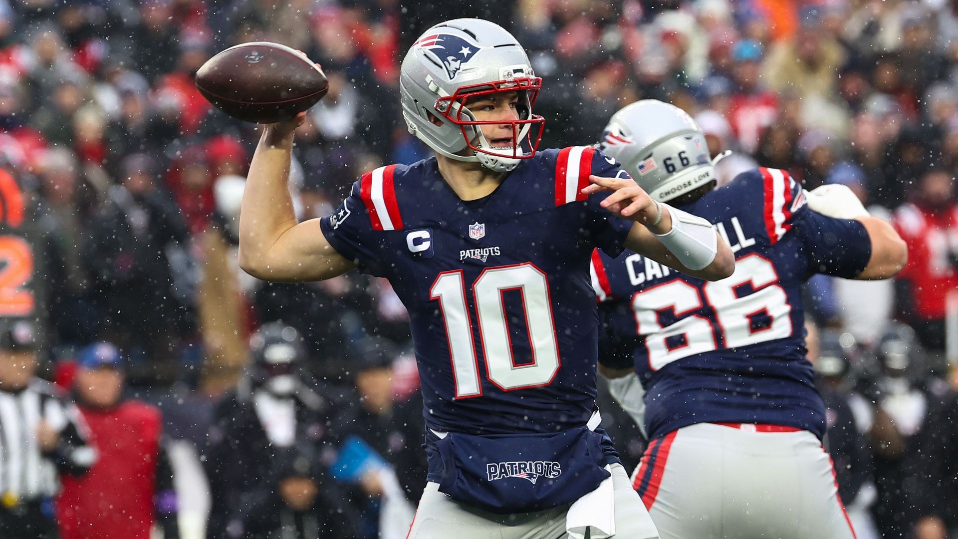 A football player in a New England Patriots uniform readies to throw the football as another player blocks a defender behind him. It's snowing and the crowd is visible in the background.