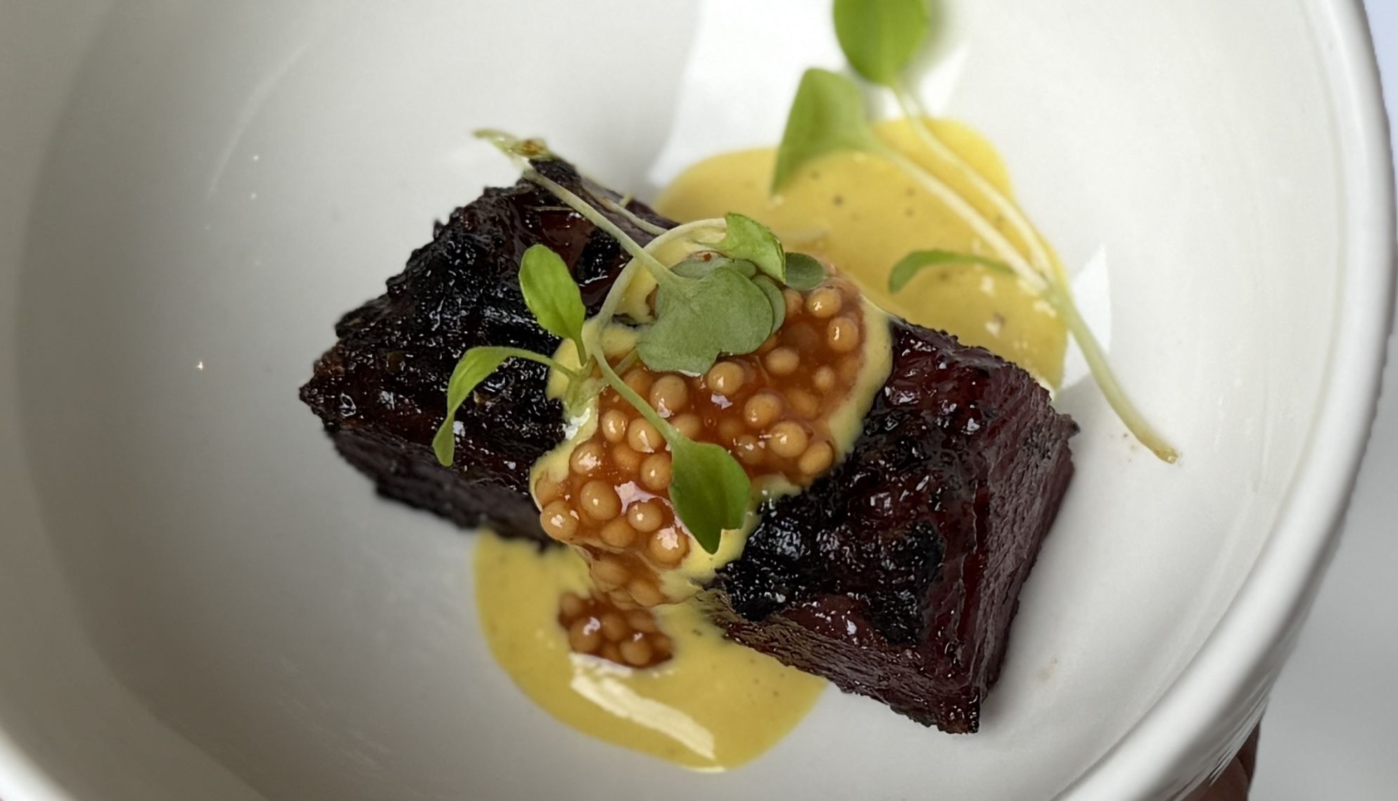 Dark, crusted beef slice topped with orange salmon roe and green microgreens, resting on a yellow sauce with a pale garnish on a white plate.