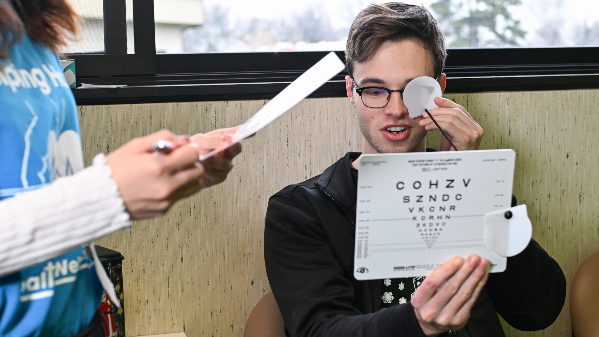 A young man undergoes a vision screening test, covering one eye while reading an eye chart, as a health care worker records the results.