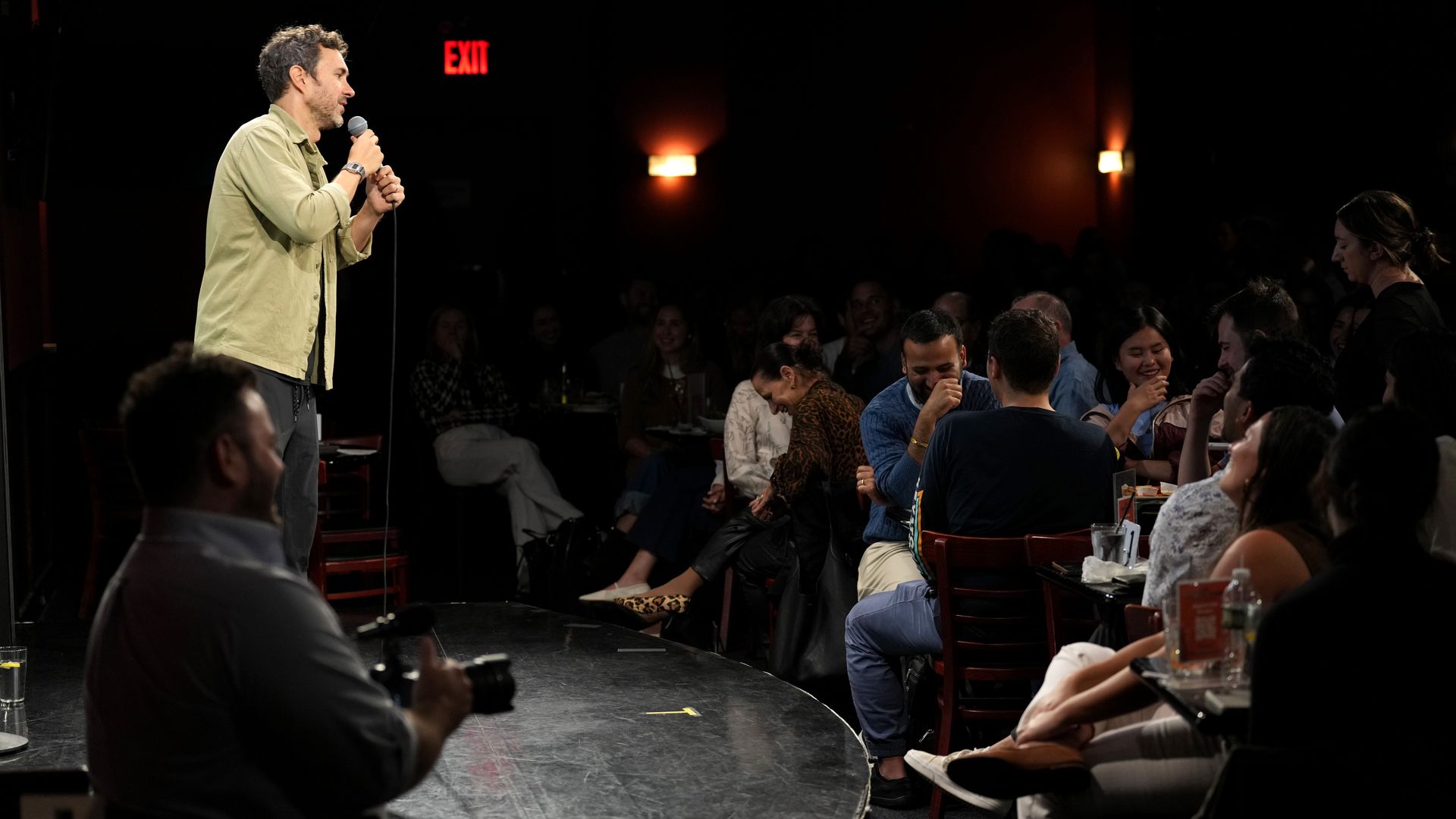 Mark Normand holds a microphone and speaks onstage in front of an audience seated at small tables.