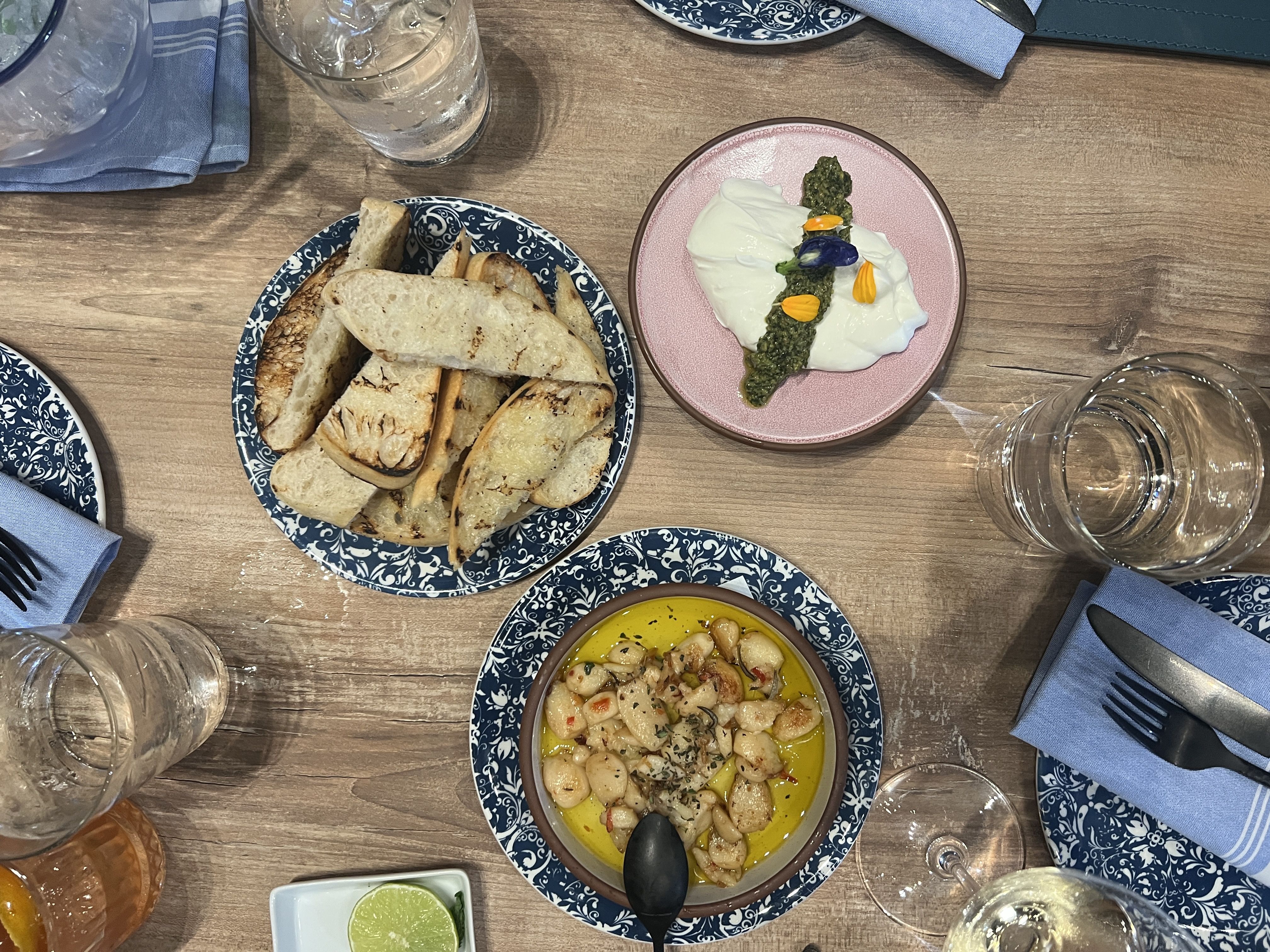 Overhead shot of a rustic table: blue patterned plates, a bread basket, pink plate with soft cheese and a green pesto stripe adorned with petals, and a yellow chickpea dish with lime wedges.