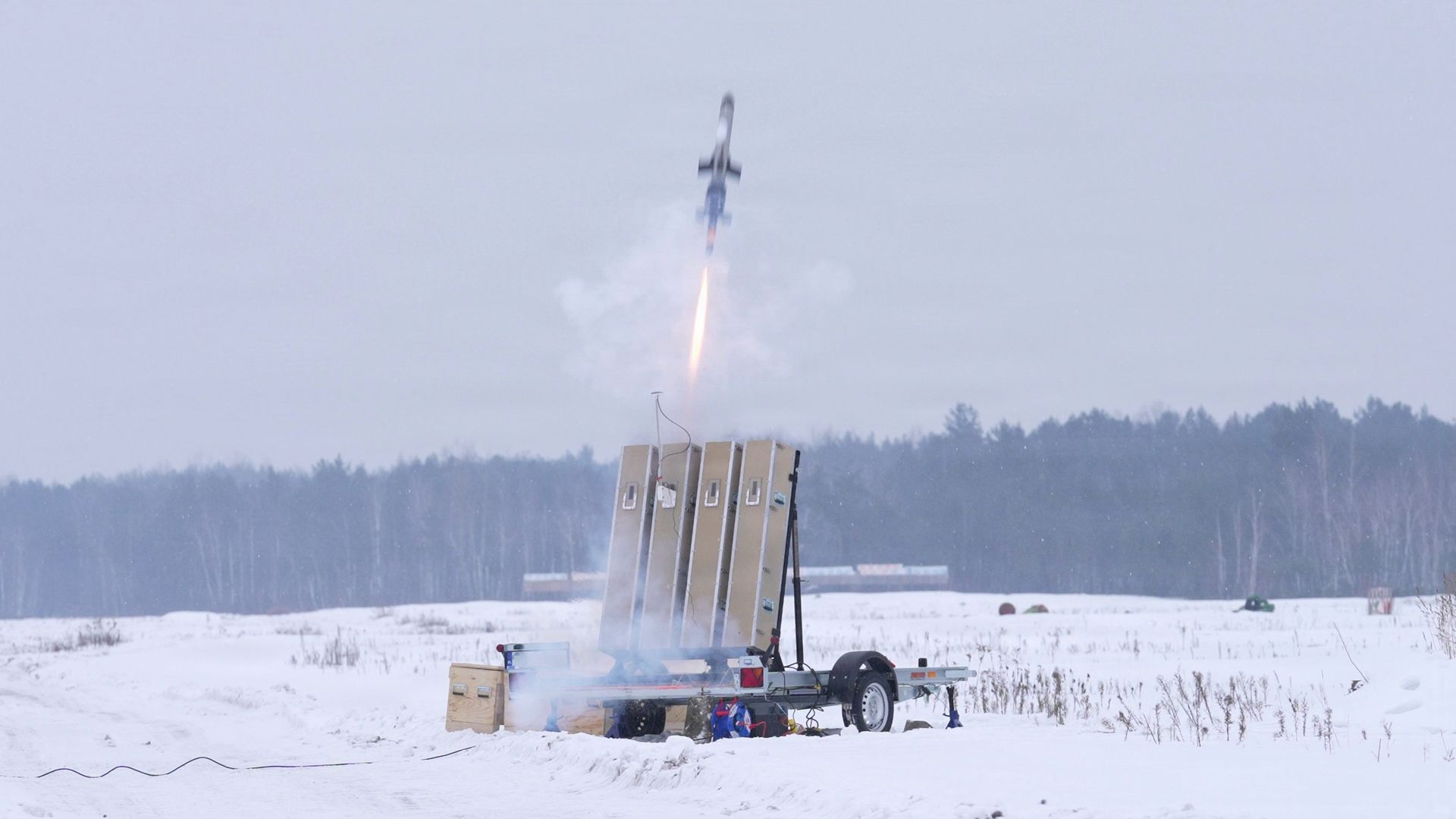 A small rocket launches vertically from a snow-covered field, spewing orange flame and white smoke. A launcher and equipment sit on a trailer; there is a trees line the horizon.