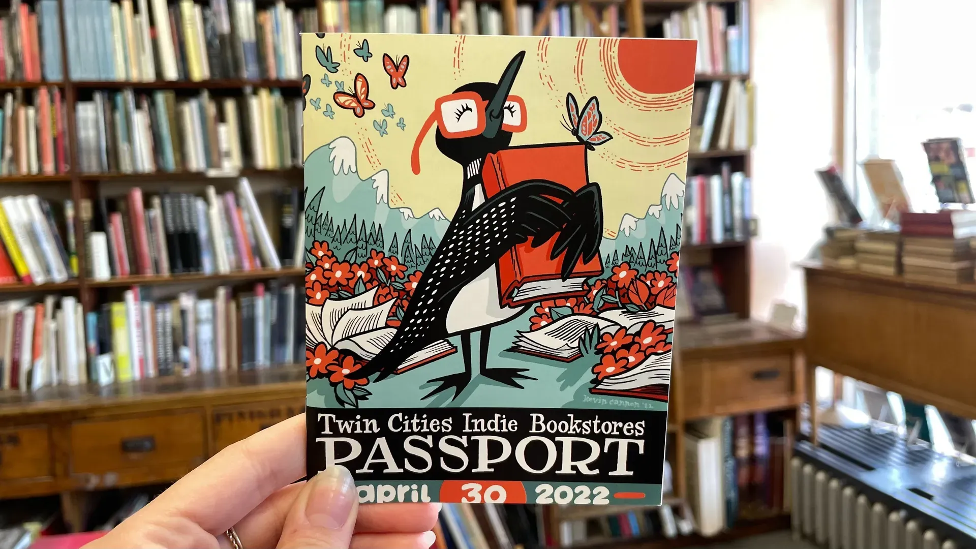 Colorful poster in a bookstore: a black bird with red glasses perched on books among red flowers and mountains, with a red sun; text reads "Twin Cities Indie Bookstores Passport" and a date.