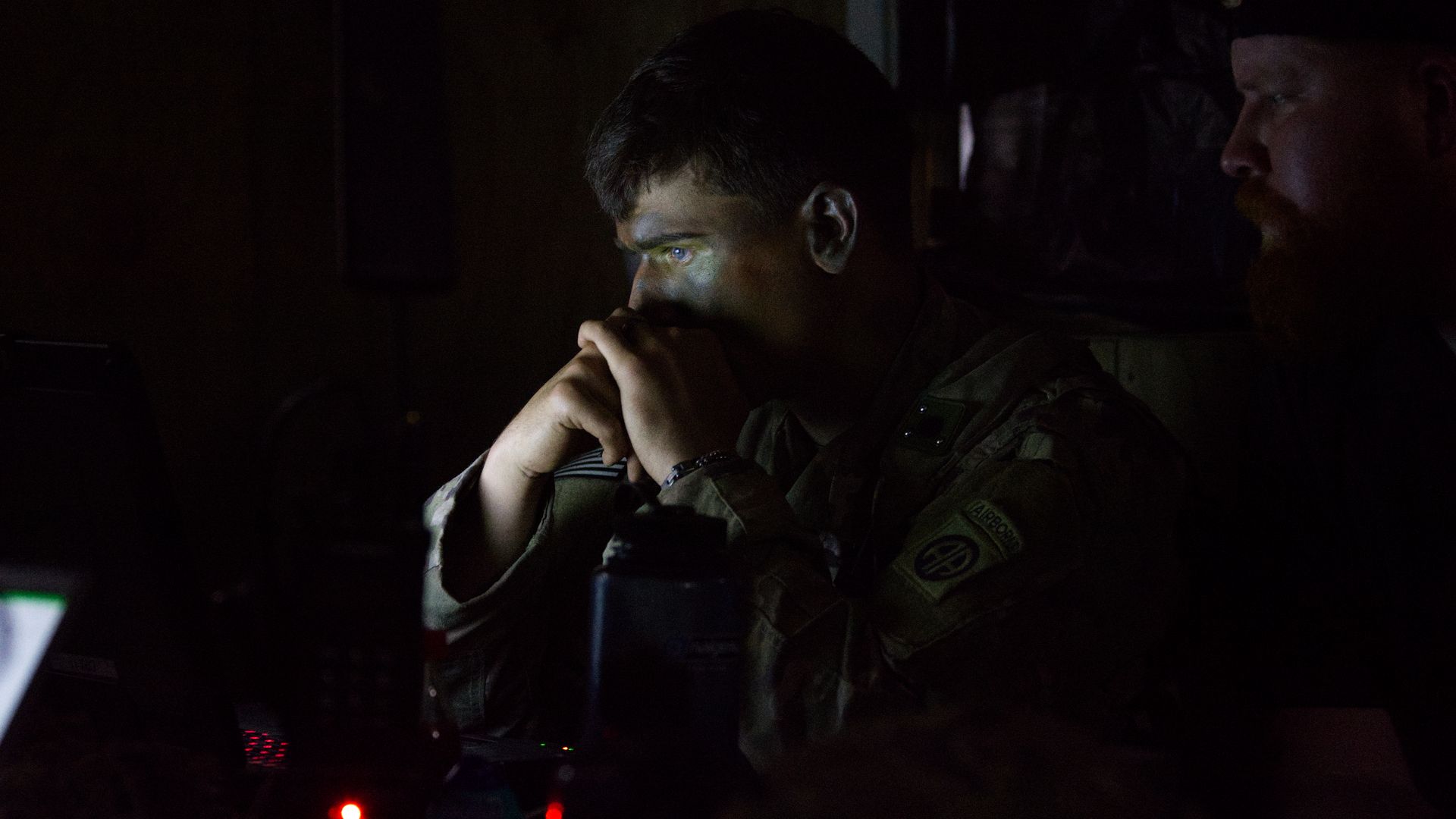 A man in camouflage military uniform sits in a dark room, hands clasped under his chin and eyes on a screen. Another bearded man sits in the shadows to his right.