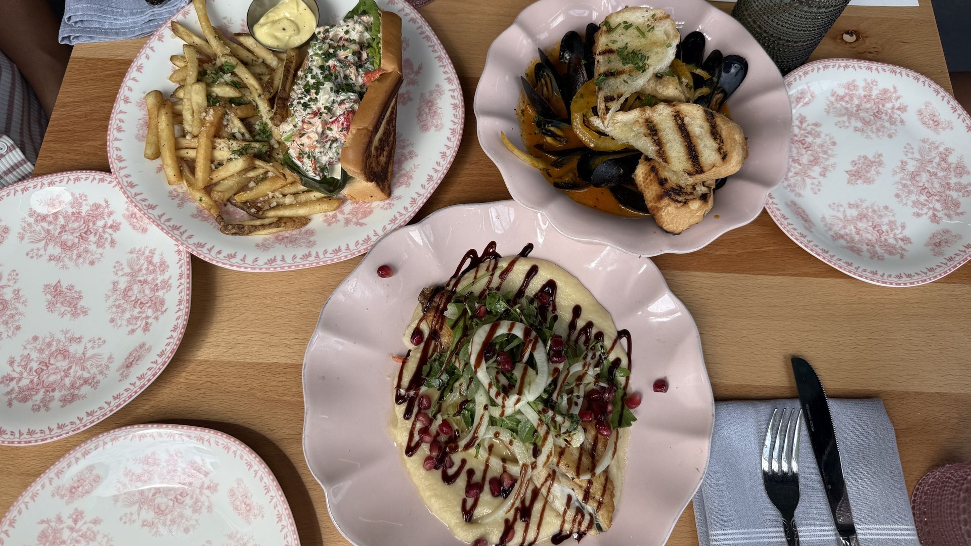 Overhead view of a wooden table with pink floral plates; left plate holds a lobster roll with fries and sauce, right plate holds mussels with grilled bread. Napkins, glassware, and a vase decorate the table.
