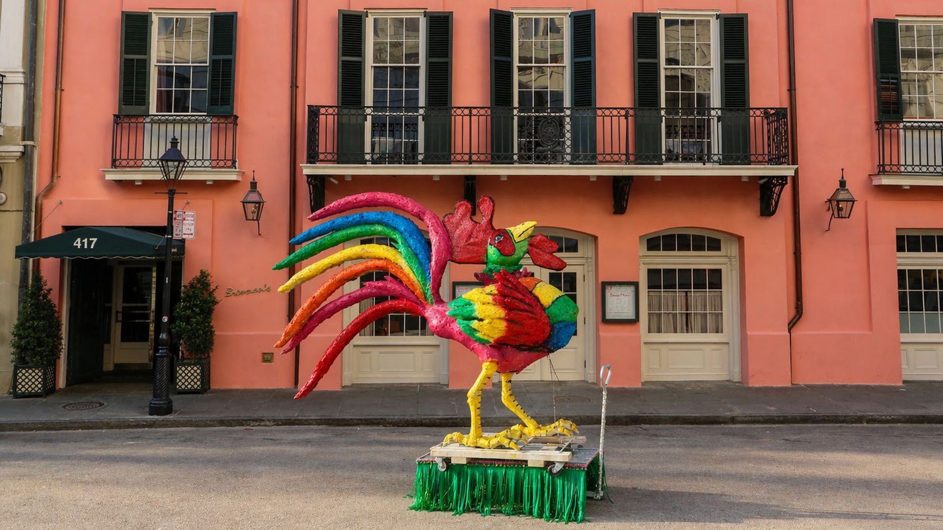Pastel pink building with black shutters and balconies; a large rainbow-colored rooster sculpture stands on a green fringed platform in the street.