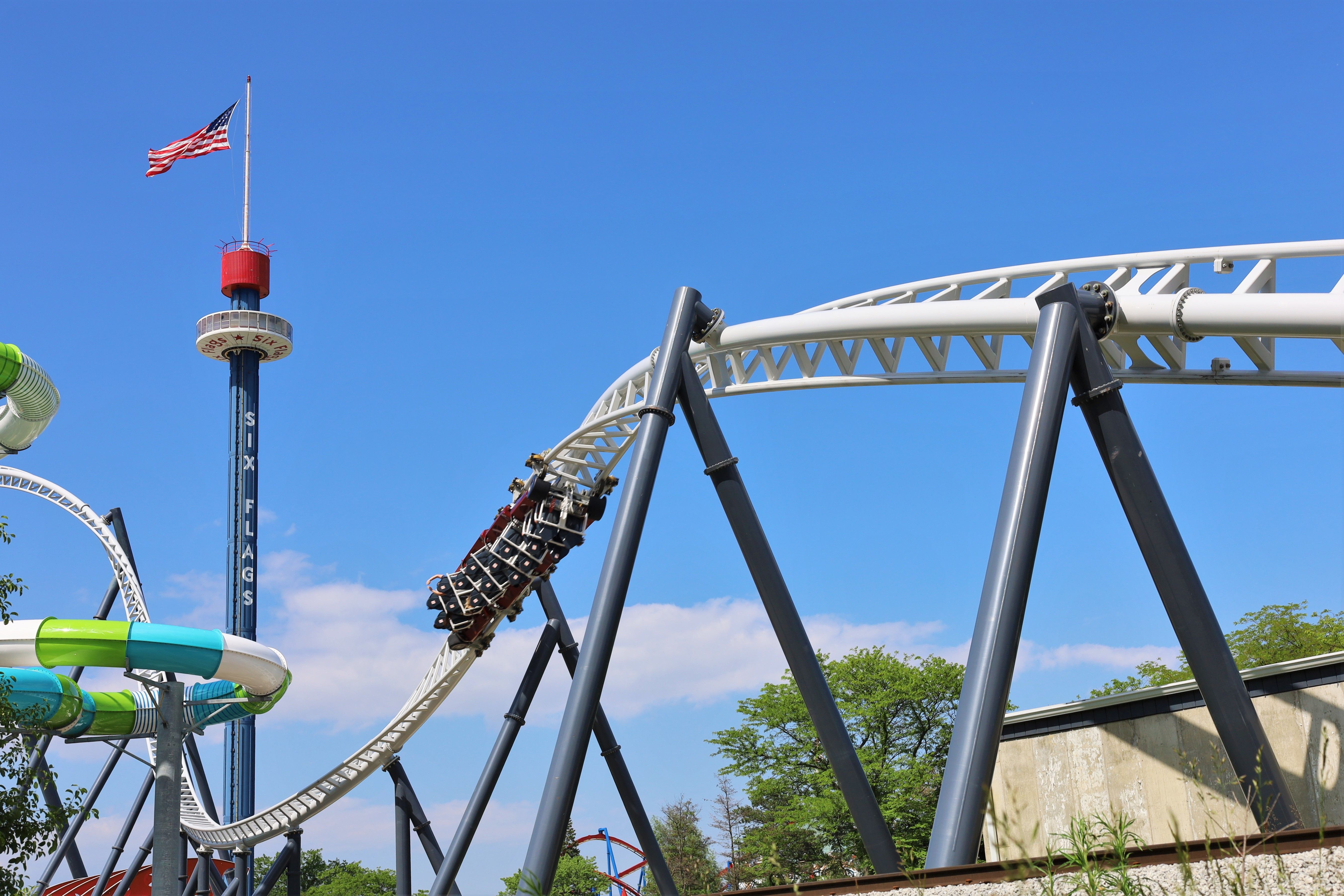 Blue-sky image of a roller coaster with white tracks and gray supports. A tall blue tower with a red top holds an American flag; curved green/blue slide structures and trees near a building.