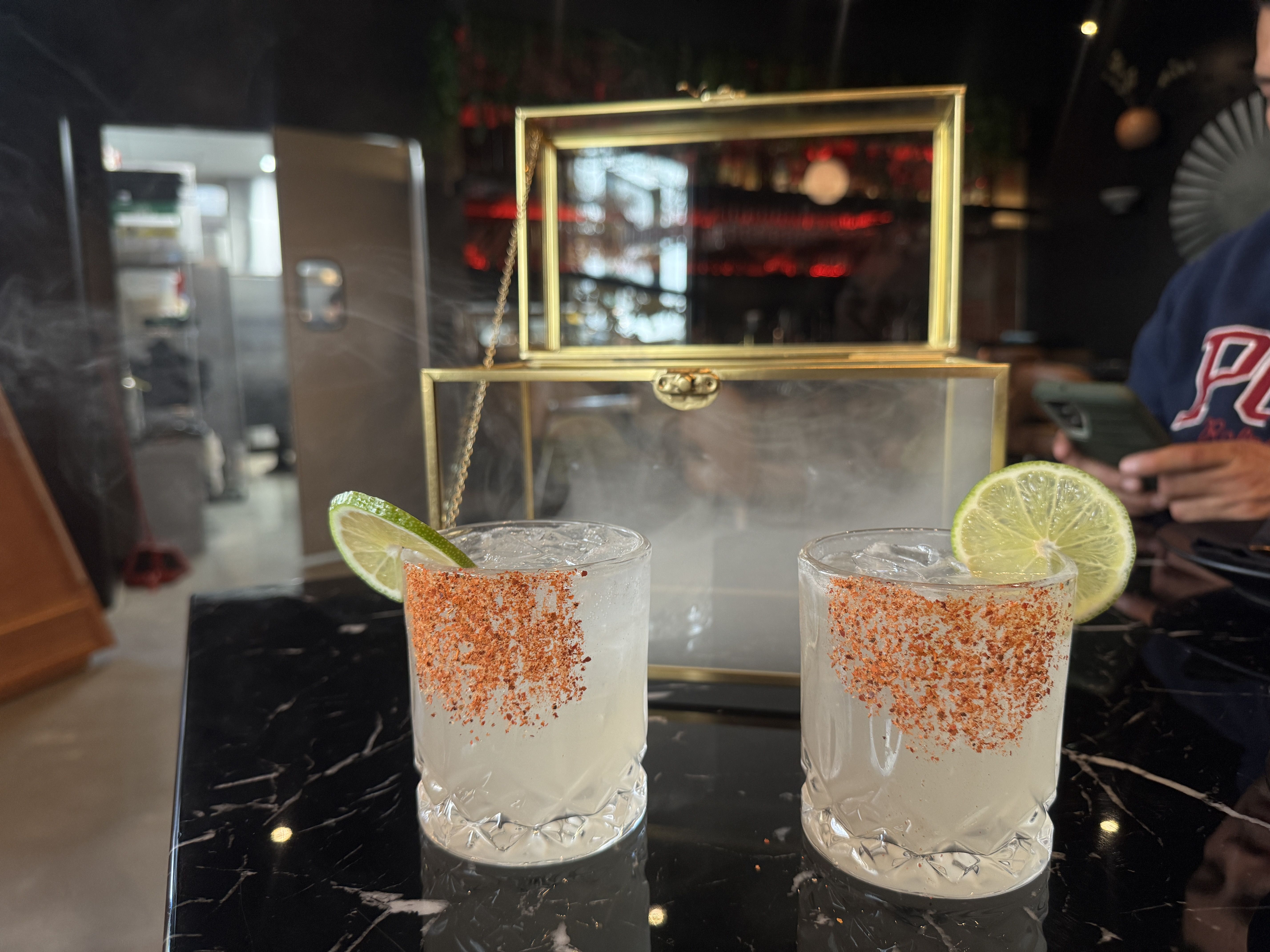 Two clear glasses with ice and salted chili rims, each garnished with a lime slice, on a black marble table with a brass and glass display case behind them.