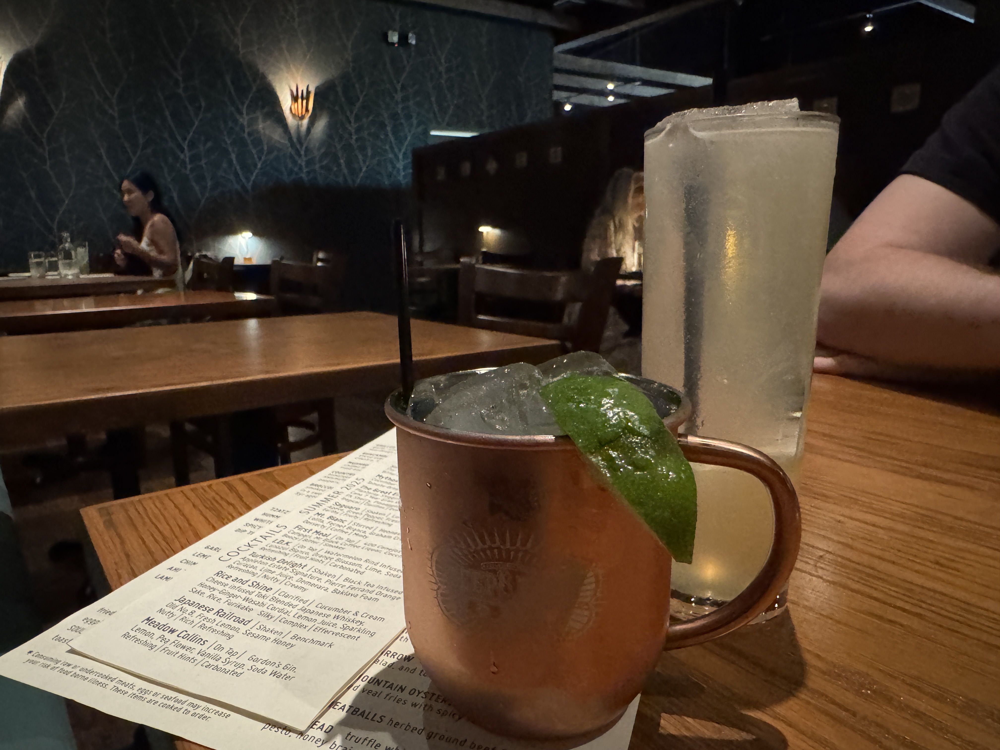 Copper mug with ice and lime wedge and a tall glass with a light-colored drink on a wooden table in a dimly lit restaurant with patterned walls and people in the background.