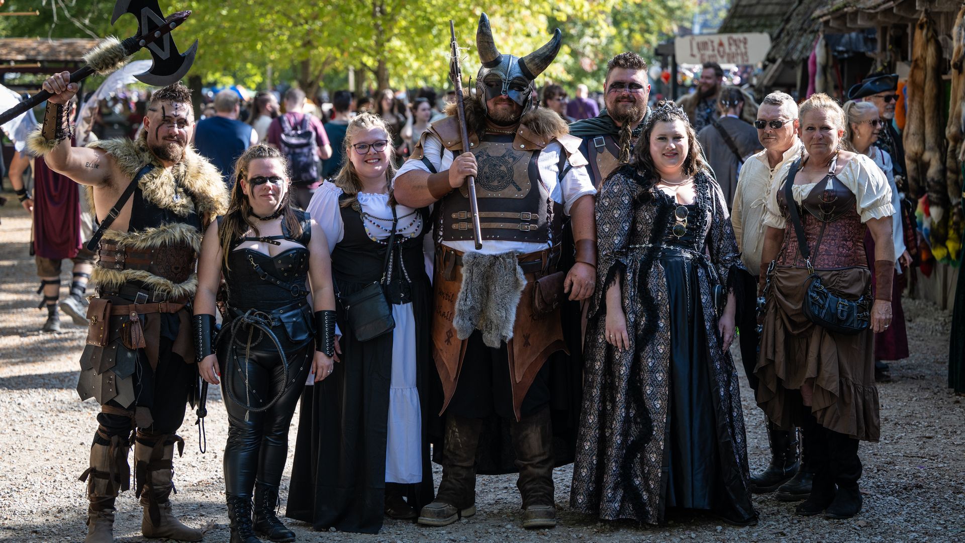 A group of festival-goers in medieval and Viking costumes pose together in a sunny outdoor market, wearing armor, fur, horns, and face paint, with a crowd and stalls in the background.